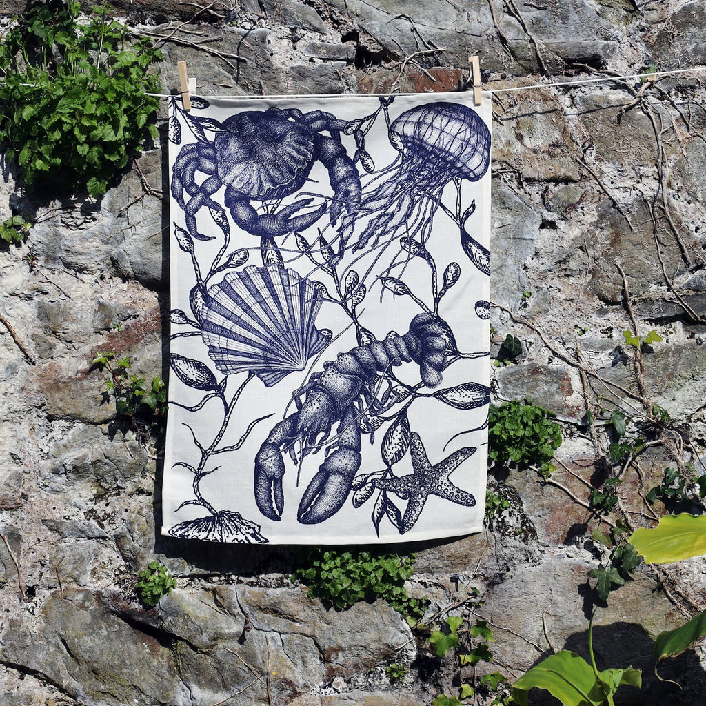 A monochrome sea-themed tea towel hangs against a rustic stone wall, secured with wooden pegs on a washing line. The design features intricate illustrations of marine life including a lobster, jellyfish, starfish, scallop shell, and seaweed, all rendered in detailed dark blue ink on a white background. Lush green plants grow around the base of the wall, adding a natural touch to the coastal-inspired scene.