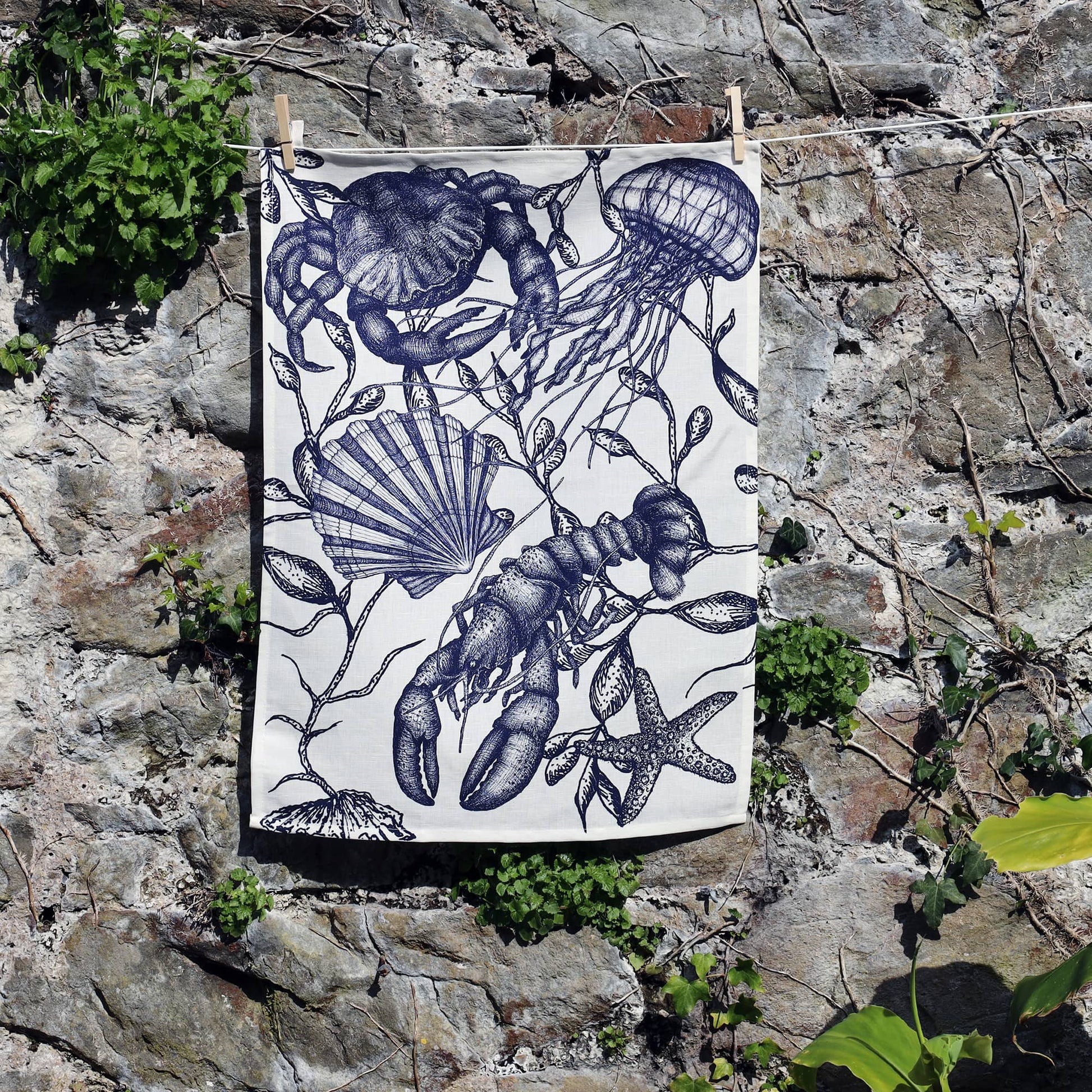 A monochrome sea-themed tea towel hangs against a rustic stone wall, secured with wooden pegs on a washing line. The design features intricate illustrations of marine life including a lobster, jellyfish, starfish, scallop shell, and seaweed, all rendered in detailed dark blue ink on a white background. Lush green plants grow around the base of the wall, adding a natural touch to the coastal-inspired scene.