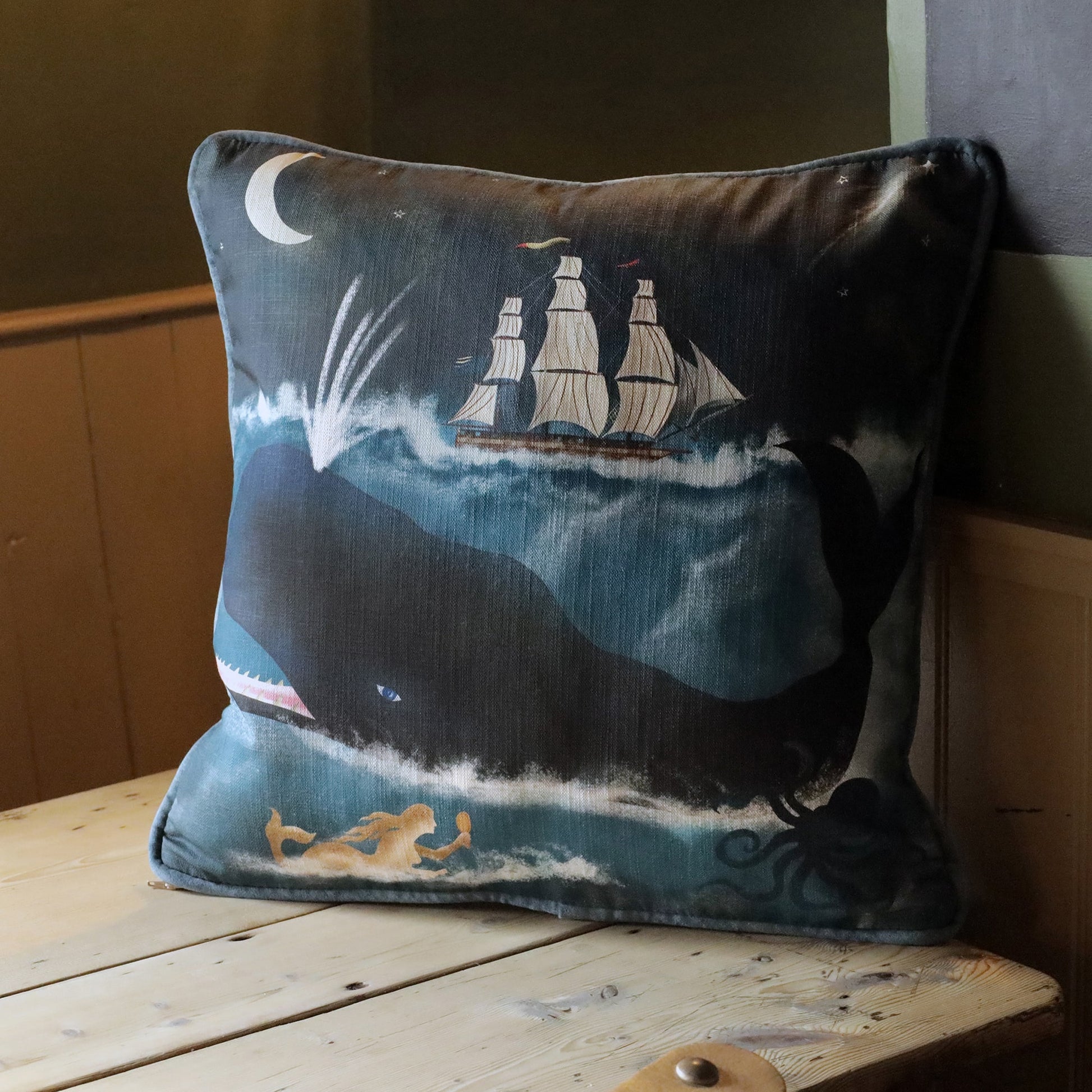A square decorative cushion featuring a nautical-themed design. The cushion showcases a whimsical scene of a large whale swimming beneath a tall ship with white sails, set against a deep blue ocean under a crescent moon. A golden mermaid swims in the lower corner, while an octopus tentacle emerges from the water. The cushion rests on a rustic wooden surface, creating a cozy, seaside-inspired atmosphere with a sense of maritime adventure and folklore.