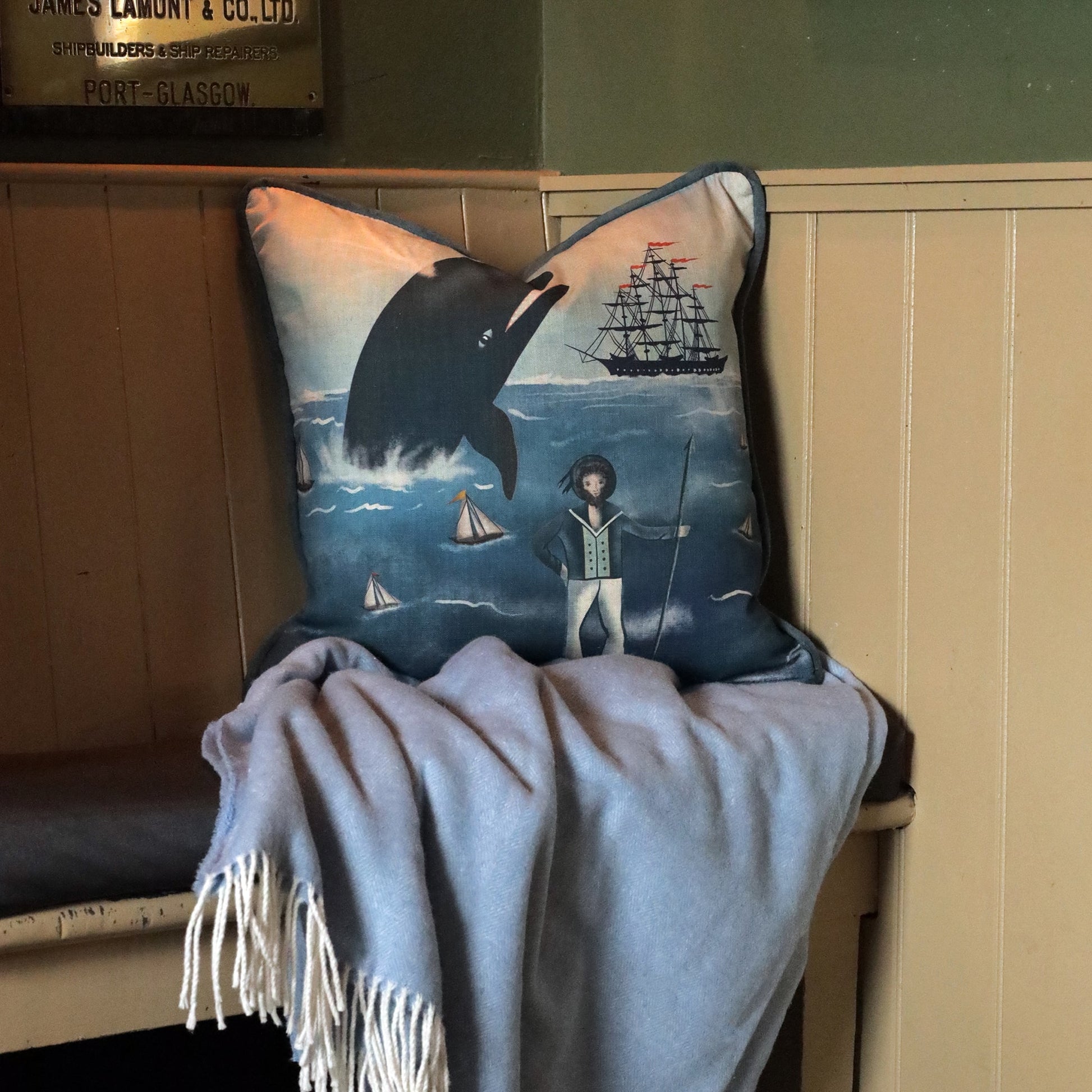 A cozy bench with a nautical-themed cushion and a soft blue blanket draped over the seat. The cushion features a whimsical design of a large whale, sailboats, and a captain standing on the shore, with a tall ship in the background. The muted tones of the cushion and blanket complement the rustic wooden paneling of the room, creating a peaceful and inviting coastal atmosphere. A vintage metal sign hangs on the wall above, adding to the maritime charm of the setting.