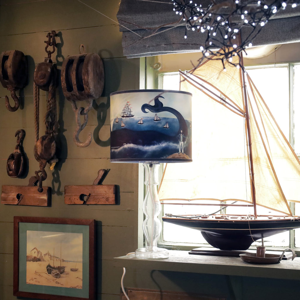 A nautical-themed room with a decorative table lamp featuring a maritime lampshade. The lampshade depicts a whimsical ocean scene with a whale, sailboats, a tall ship, and a captain standing on the shore. The lamp is placed on a windowsill beside a large model sailboat with cream-colored sails. The room is decorated with rustic maritime tools and pulleys hanging on the wall, alongside a framed painting of boats on a beach. Soft lighting and warm wood accents create a cozy, seaside atmosphere, enhanced by a 