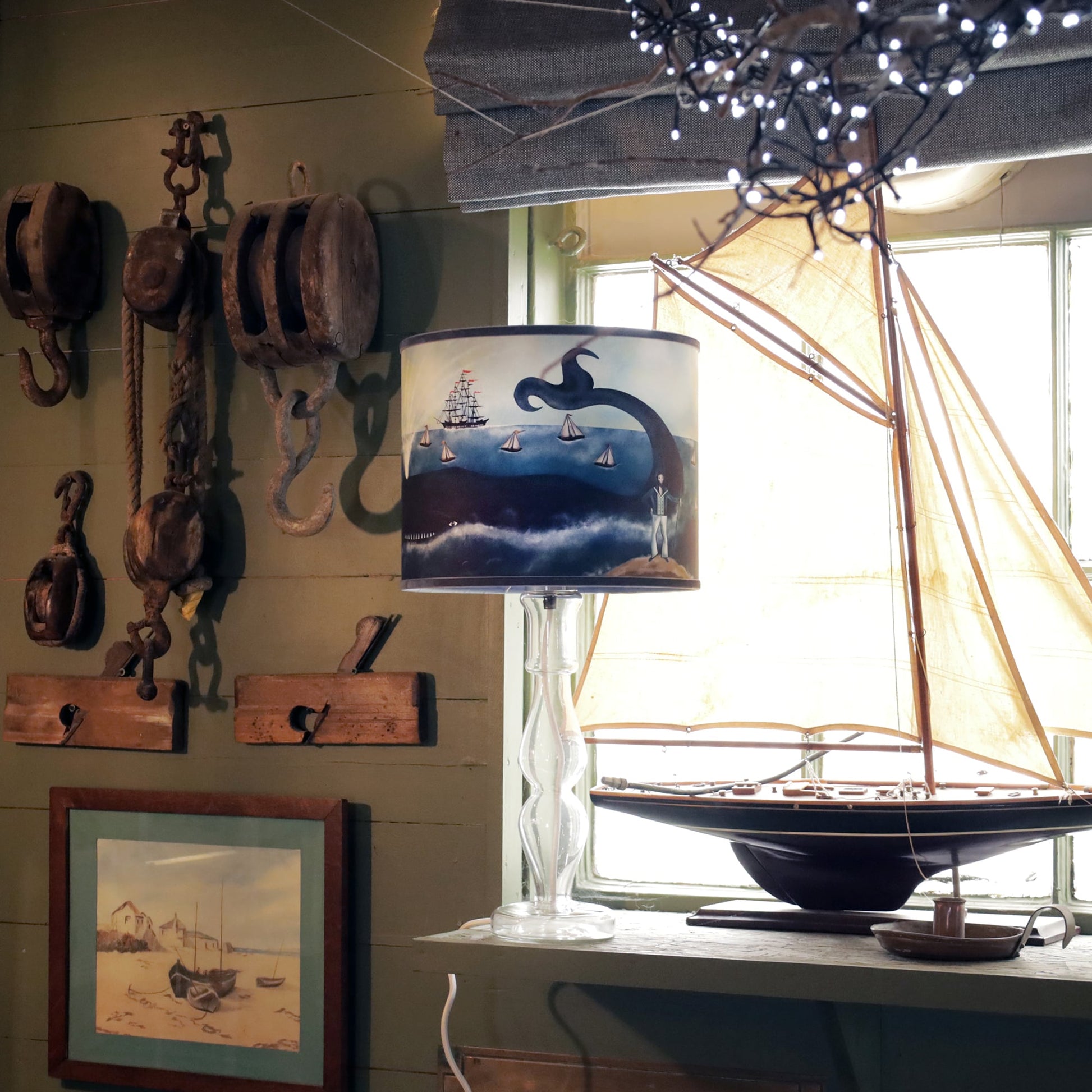 A nautical-themed room with a decorative table lamp featuring a maritime lampshade. The lampshade depicts a whimsical ocean scene with a whale, sailboats, a tall ship, and a captain standing on the shore. The lamp is placed on a windowsill beside a large model sailboat with cream-colored sails. The room is decorated with rustic maritime tools and pulleys hanging on the wall, alongside a framed painting of boats on a beach. Soft lighting and warm wood accents create a cozy, seaside atmosphere, enhanced by a 