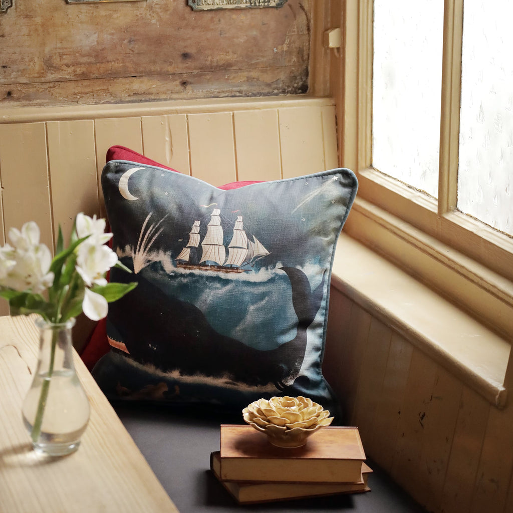 A cozy window seat featuring a nautical-themed cushion with an illustration of a whale and a tall ship under a crescent moon. The cushion rests against a red pillow, adding a pop of color to the serene space. A small stack of vintage books and a decorative ceramic flower sit on the bench beside the cushion. A vase of white flowers on the nearby table adds a touch of freshness. Soft light filters through the window, enhancing the peaceful, seaside-inspired ambiance of the room.
