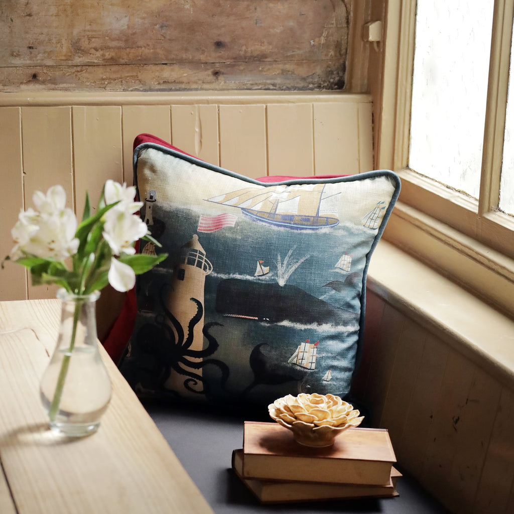  A cozy window nook featuring a nautical-themed cushion with an illustration of a large whale, a tall ship, and a crescent moon. The cushion rests against a red pillow, adding a pop of color to the serene space. A small stack of vintage books and a decorative ceramic flower sit on the bench beside the cushion. A vase with white flowers on the nearby table adds a fresh touch. Soft natural light streams through the window, creating a warm and inviting seaside-inspired atmosphere.