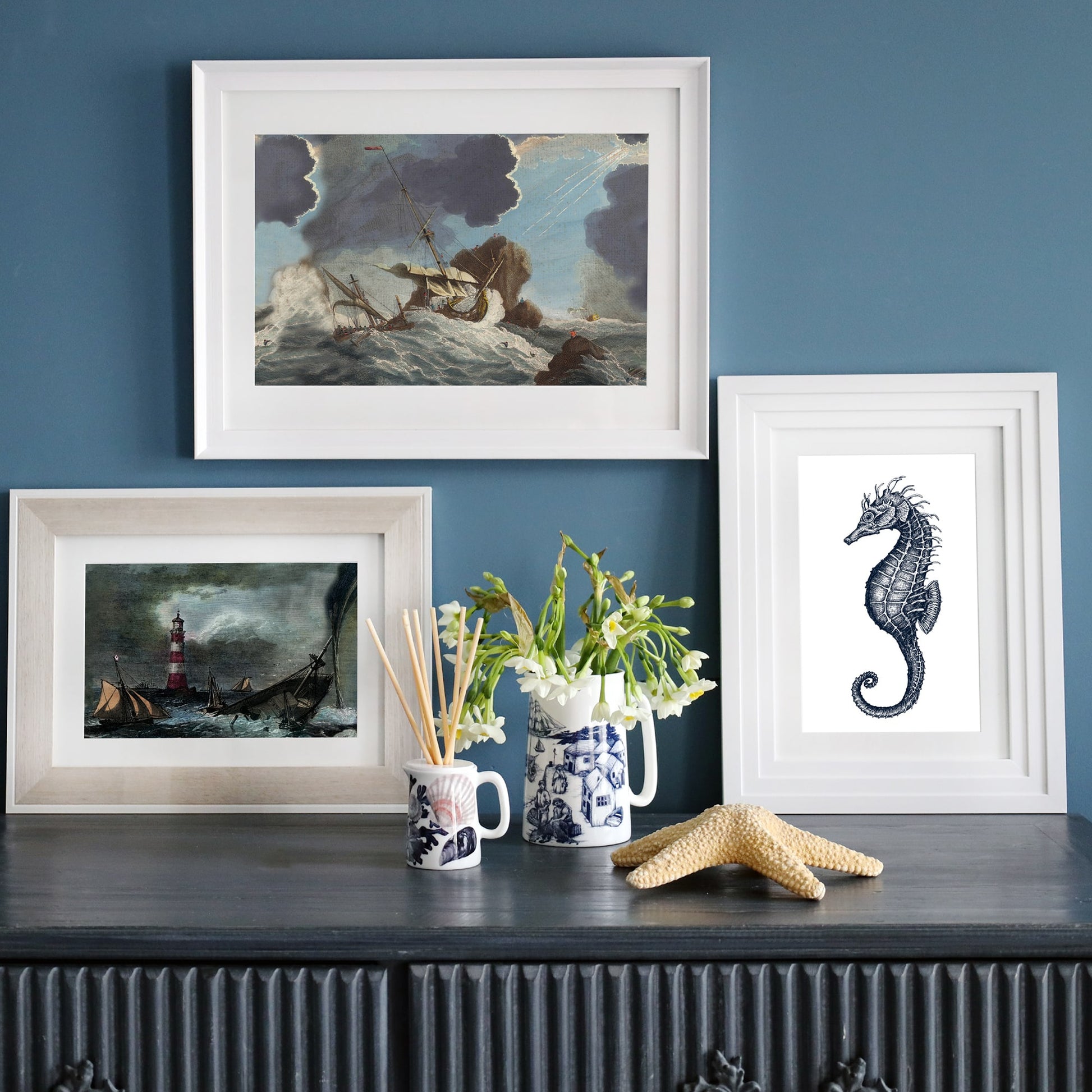A nautical-themed display featuring three framed artworks on a dark wooden sideboard against a deep blue wall. The top frame showcases a dramatic shipwreck scene with stormy skies, the bottom left frame features a lighthouse with ships in turbulent waters, and the right frame displays a detailed black-and-white seahorse illustration. The arrangement is complemented by a ceramic jug with white flowers, a reed diffuser in a maritime-patterned mug, and a textured starfish.