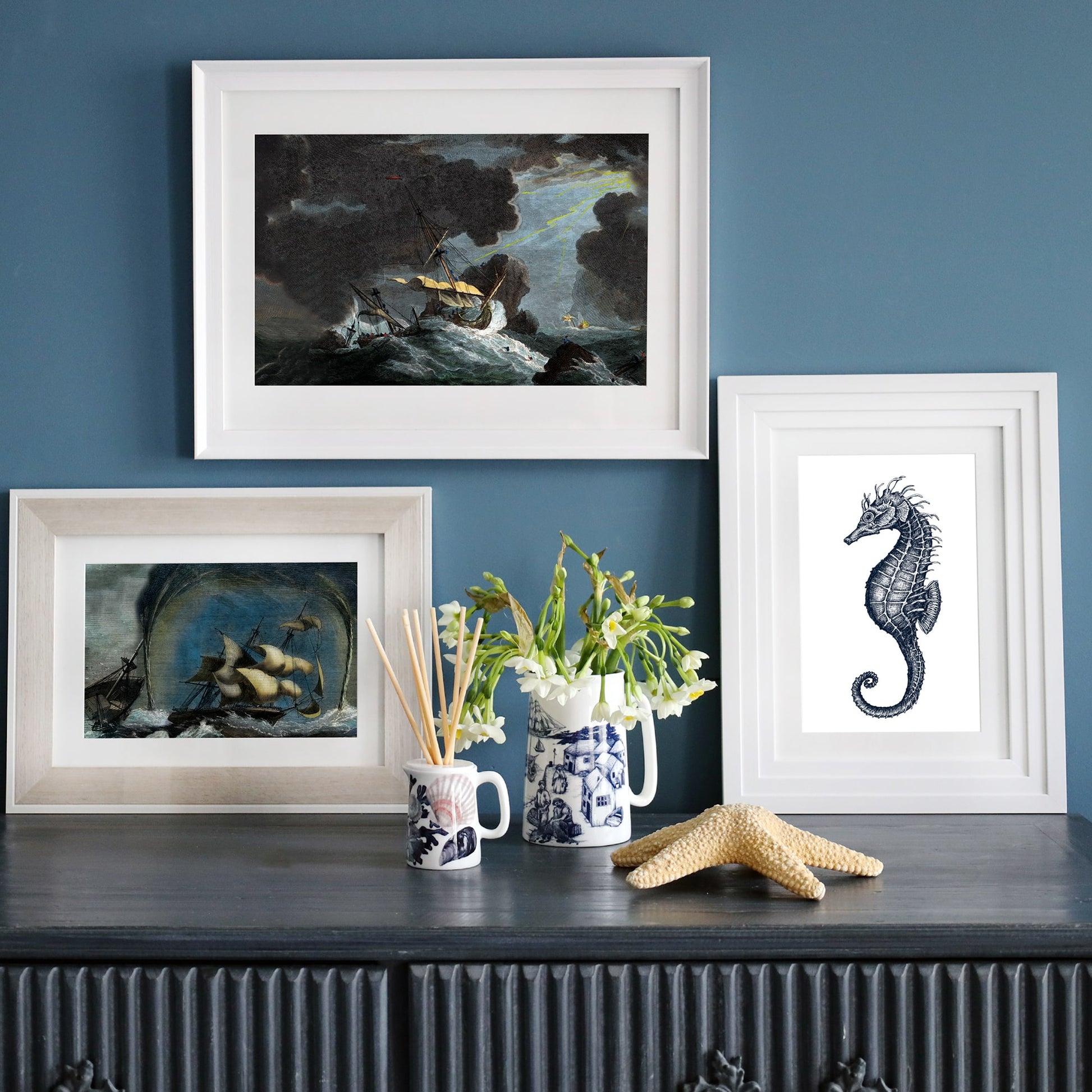 Three framed nautical prints on a dark blue wall, featuring a stormy shipwreck, a sailing ship in a whirlwind, and a detailed seahorse illustration, styled on a sideboard with coastal-inspired decor