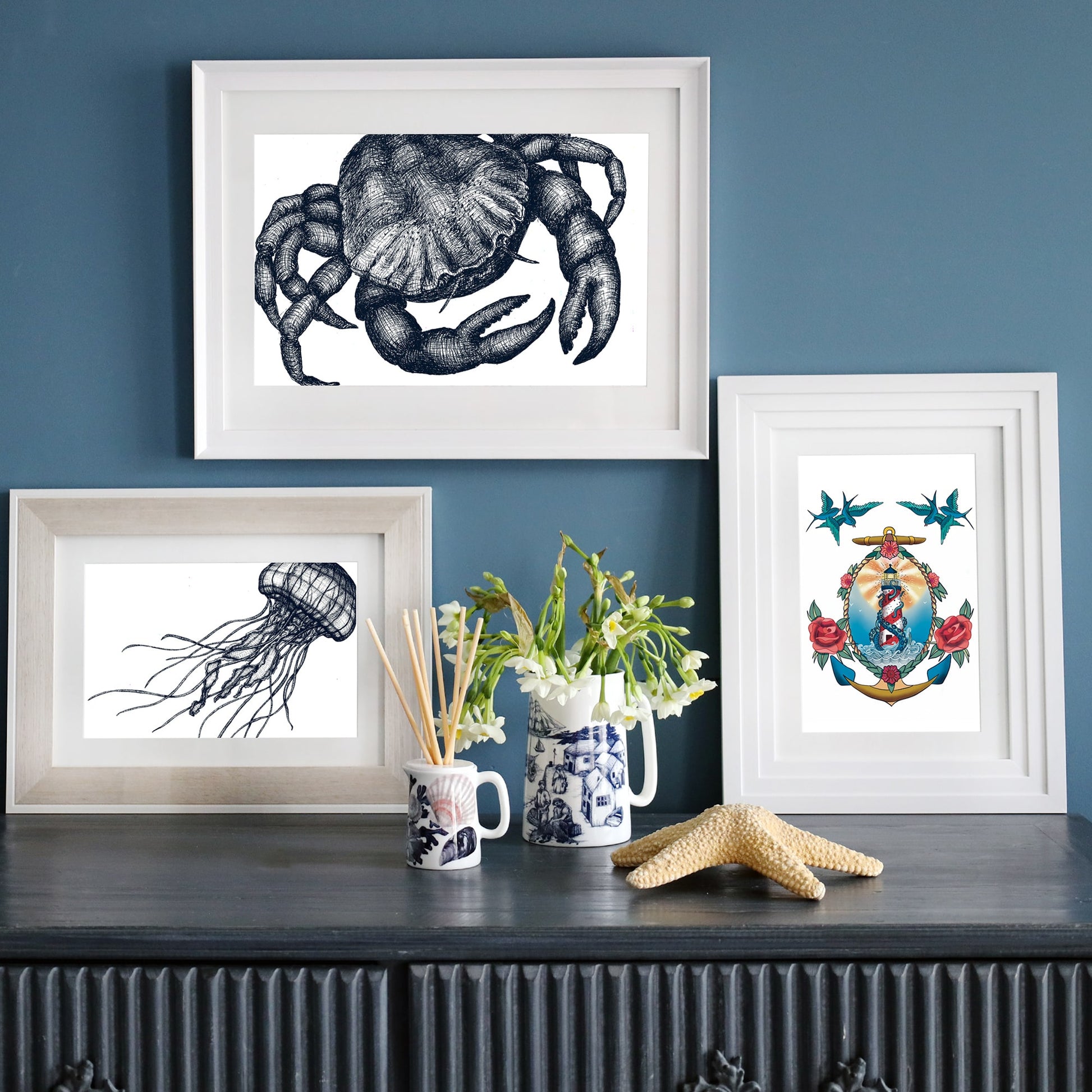 Three framed nautical prints displayed on a dark wooden surface against a deep blue wall. The top frame features a detailed black-and-white crab illustration, the bottom left frame showcases a jellyfish with flowing tentacles, and the right frame contains a colorful lighthouse wrapped in a kraken’s tentacles, surrounded by roses and swallows. A starfish, a vase with flowers, and decorative ceramic pieces complement the arrangement.