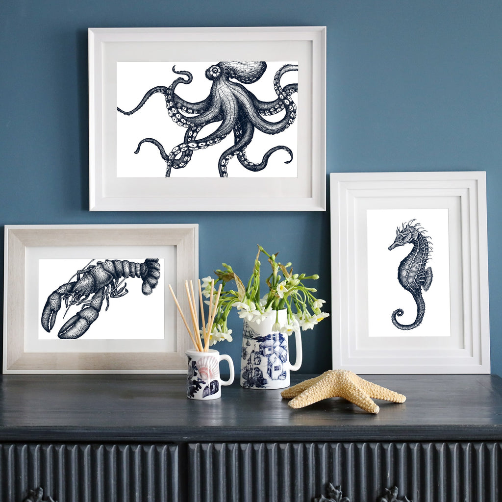 A set of three framed blue-and-white marine life illustrations—a detailed octopus, a lobster, and a seahorse—displayed on a dark blue wall. Below, a dark sideboard is decorated with a starfish, a vase of white flowers, ceramic mugs, and reed diffusers.