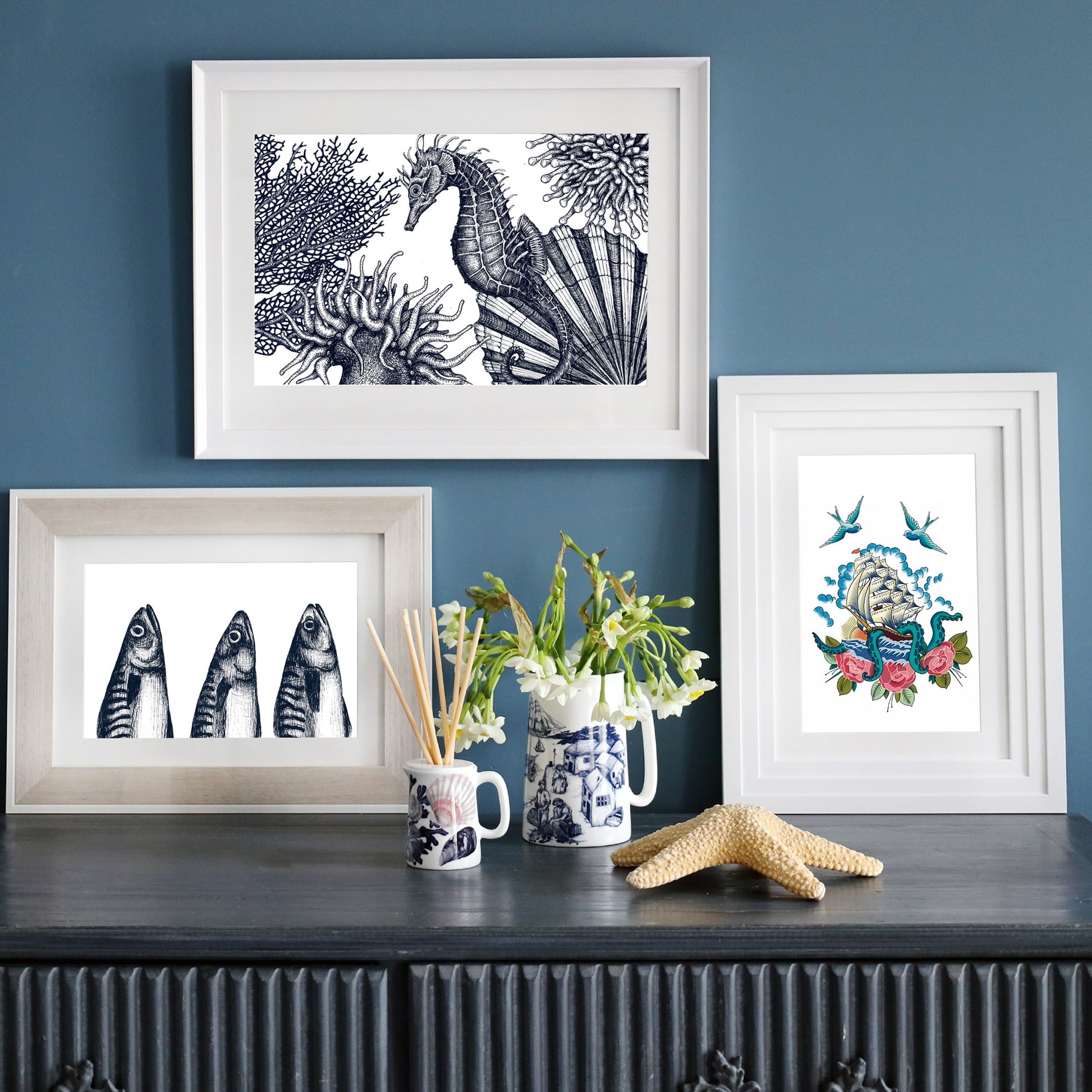 A collection of three framed nautical prints on a dark blue wall. The top print features a detailed black-and-white seahorse scene, the bottom left shows three mackerel heads, and the right print presents a colorful tattoo-style illustration of a sailing ship with swallows, roses, and waves.