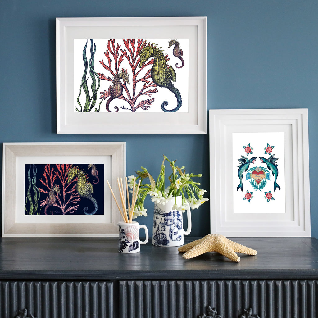 A styled interior with three framed prints against a deep blue wall: two featuring seahorses with coral (one on white, one on navy) and one with swallows and a heart. Displayed on a dark wooden sideboard with a ceramic jug of flowers, a starfish, and nautical-themed decor.