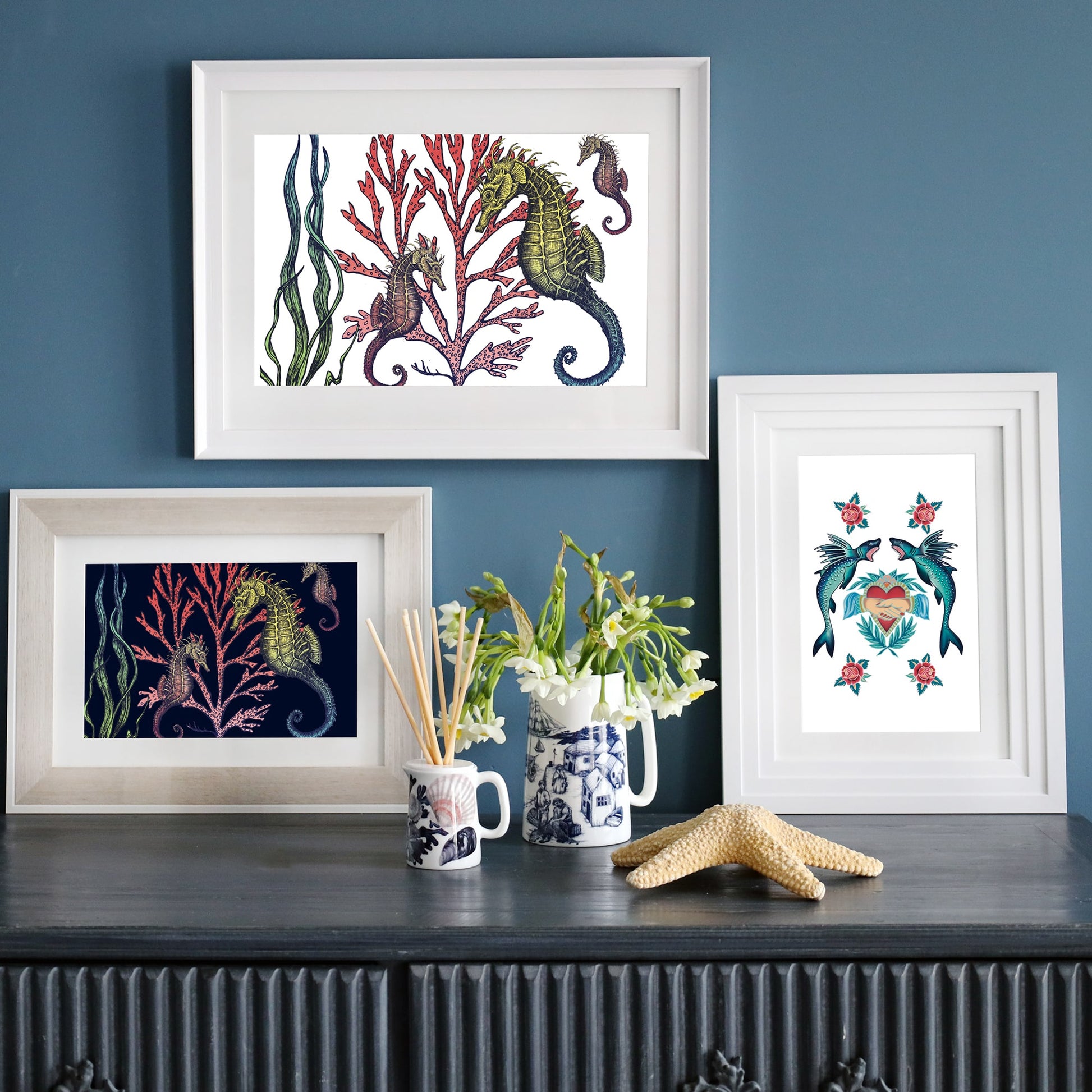 A dark sideboard against a deep blue wall displays three framed nautical prints—two featuring seahorses among coral and one with stylized sharks and a heart motif. Decor accents include a starfish, a vase with white flowers, blue and white ceramic mugs, and a reed diffuser.
