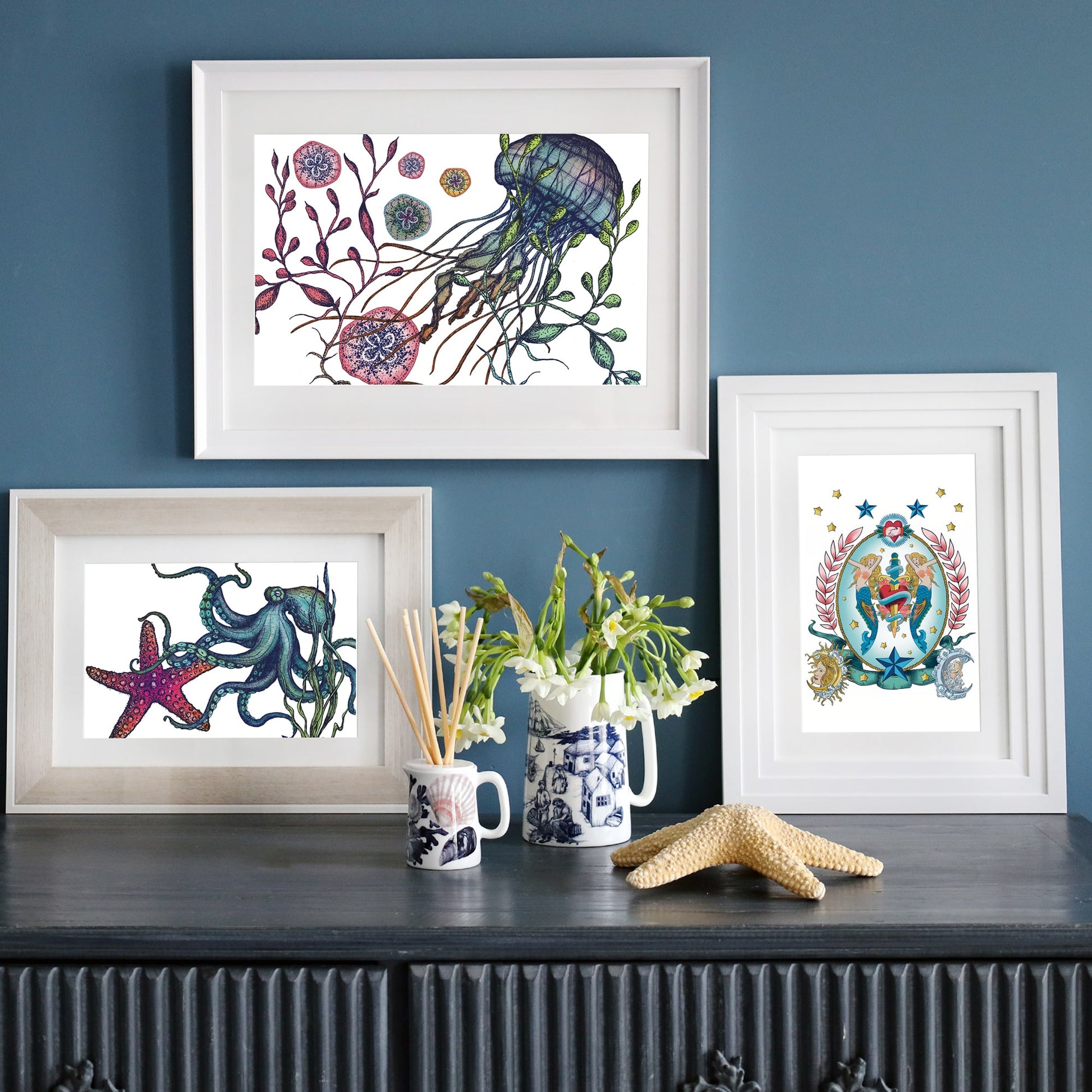 A dark sideboard against a deep blue wall features three framed ocean-themed prints—one with a jellyfish, another with an octopus and starfish, and a third with a mermaid in a decorative frame. Decor includes a starfish, a vase of white flowers, blue and white ceramic mugs, and a reed diffuser.