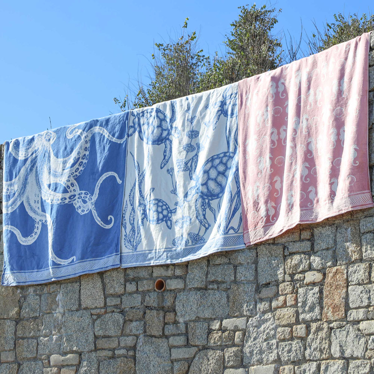 Three patterned reversible throws hanging outdoors on a line above a stone wall under a blue sky. Two towels are white with blue sea-themed prints (octopus and turtle), and one towel is pink with white seahorse prints.