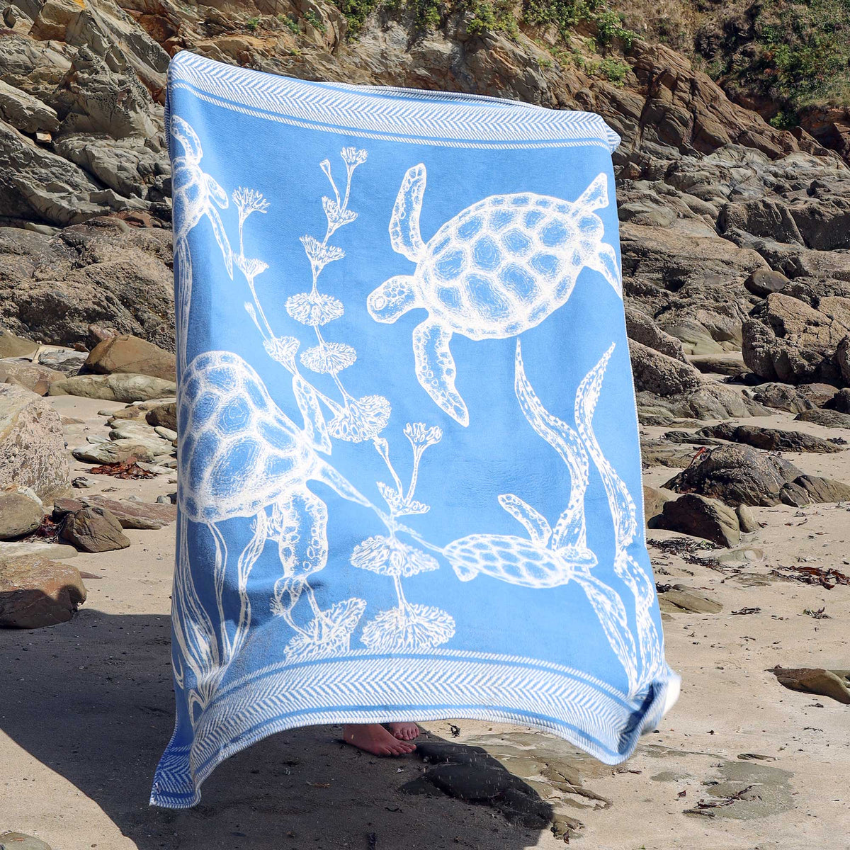 A light blue reversible throw with white sea turtle and sea plant illustrations, draped over a person standing on a rocky beach.