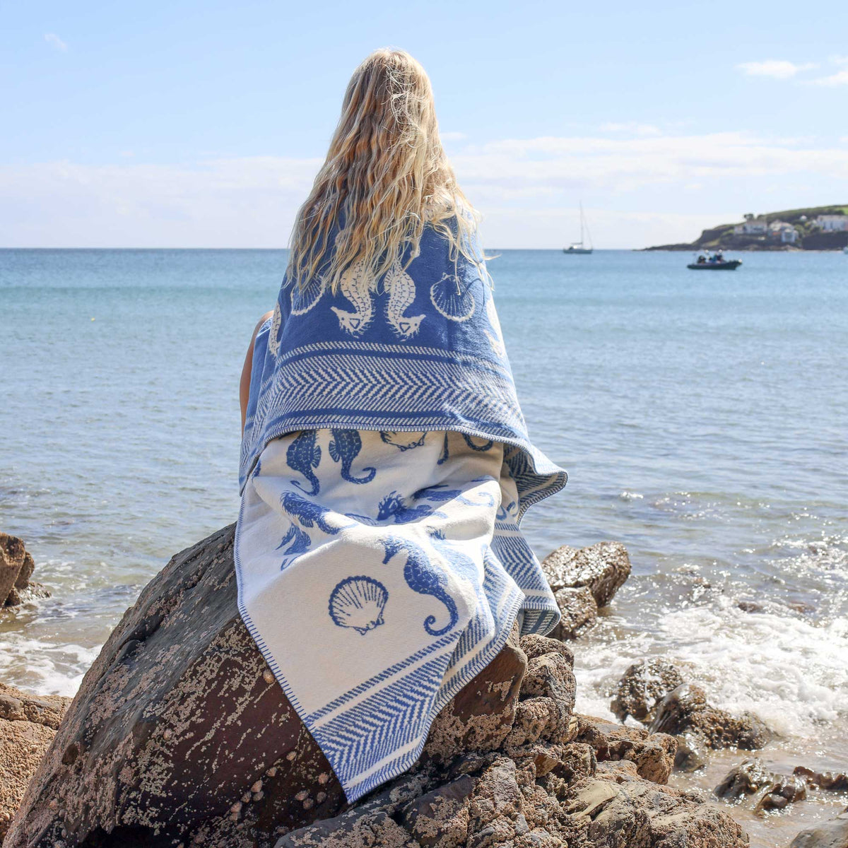 A woman sits on a rock by the ocean, wrapped in a blue and white patterned blanket featuring seahorse and shell motifs. The background shows calm blue water, distant boats, and a shoreline under a bright sky.