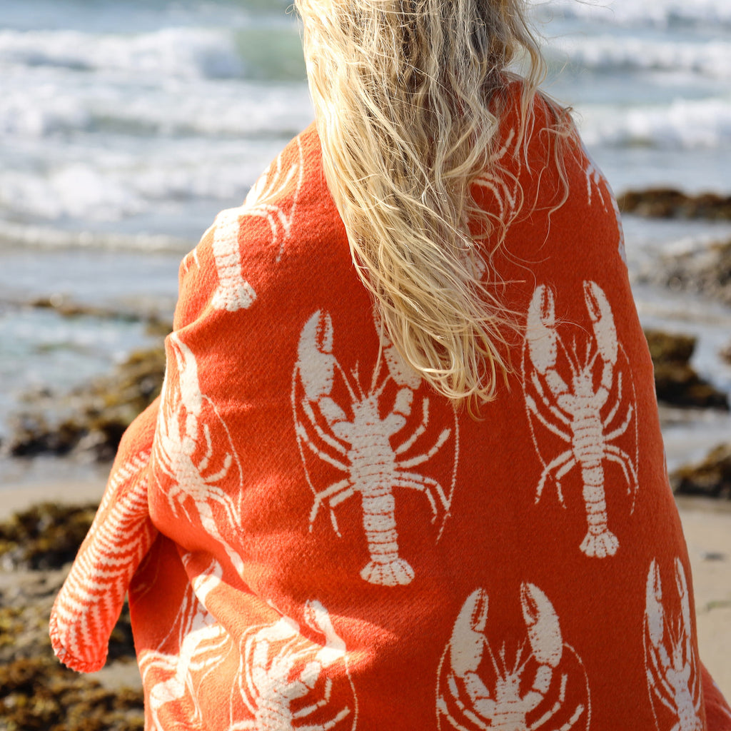 Person wearing an orange blanket with white lobster patterns on a beach.