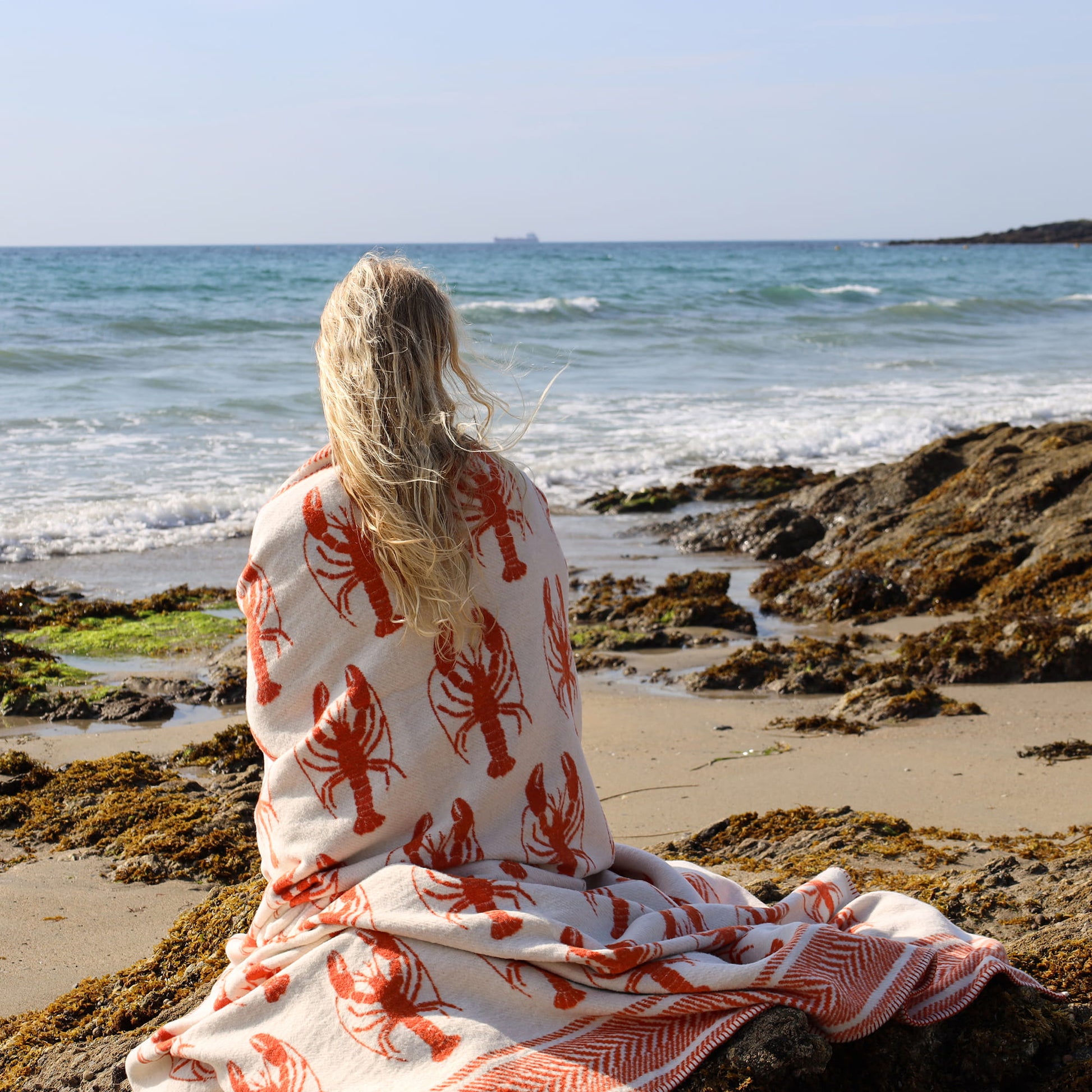 Person sitting on a rocky beach wearing a blanket with red lobster patterns.