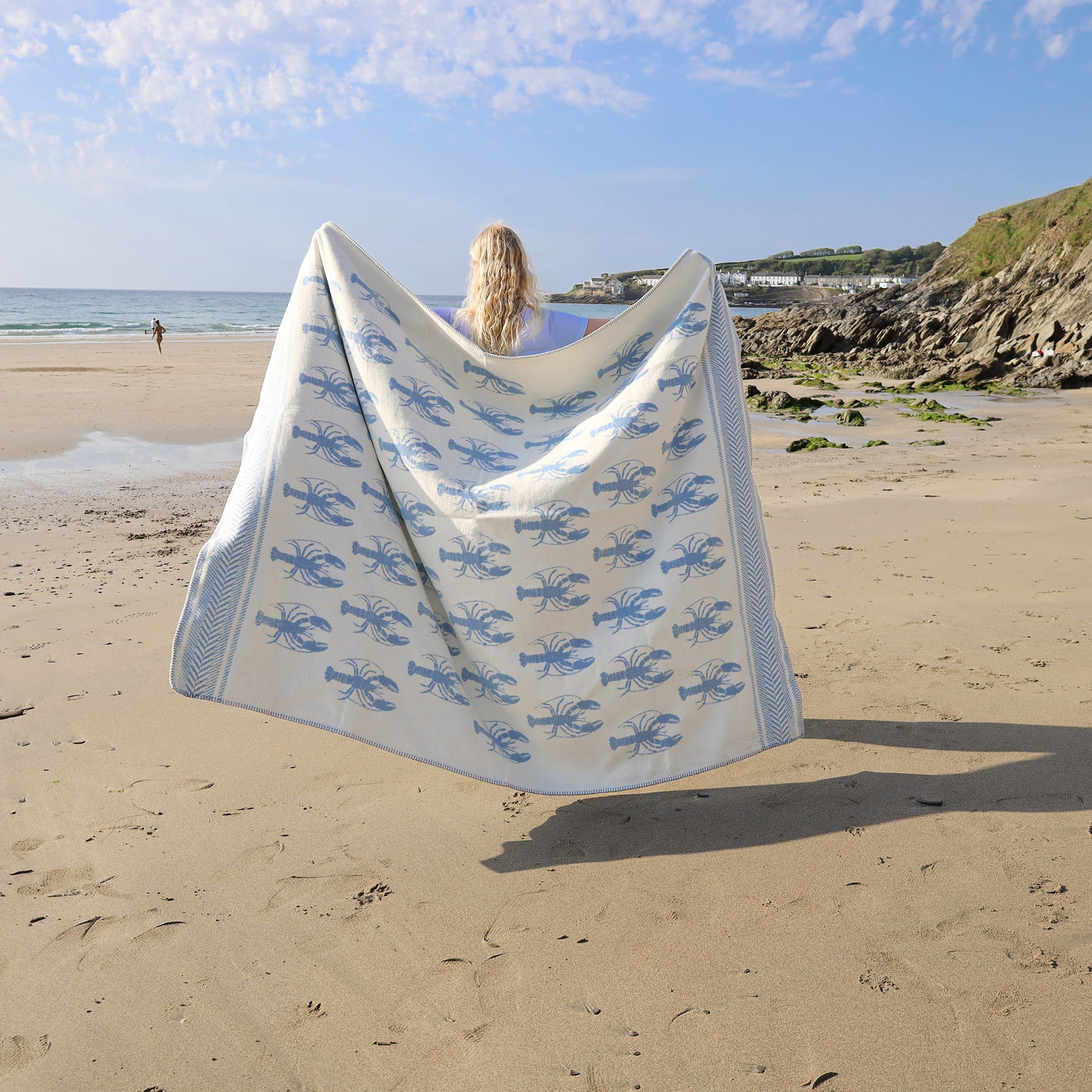 Person holding a beach towel with blue lobster pattern on a sandy beach.