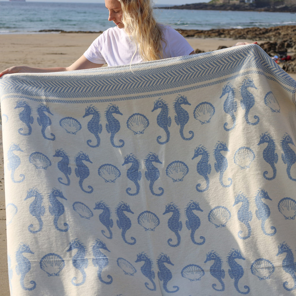 Person holding a blanket with blue seahorse patterns on a beach.