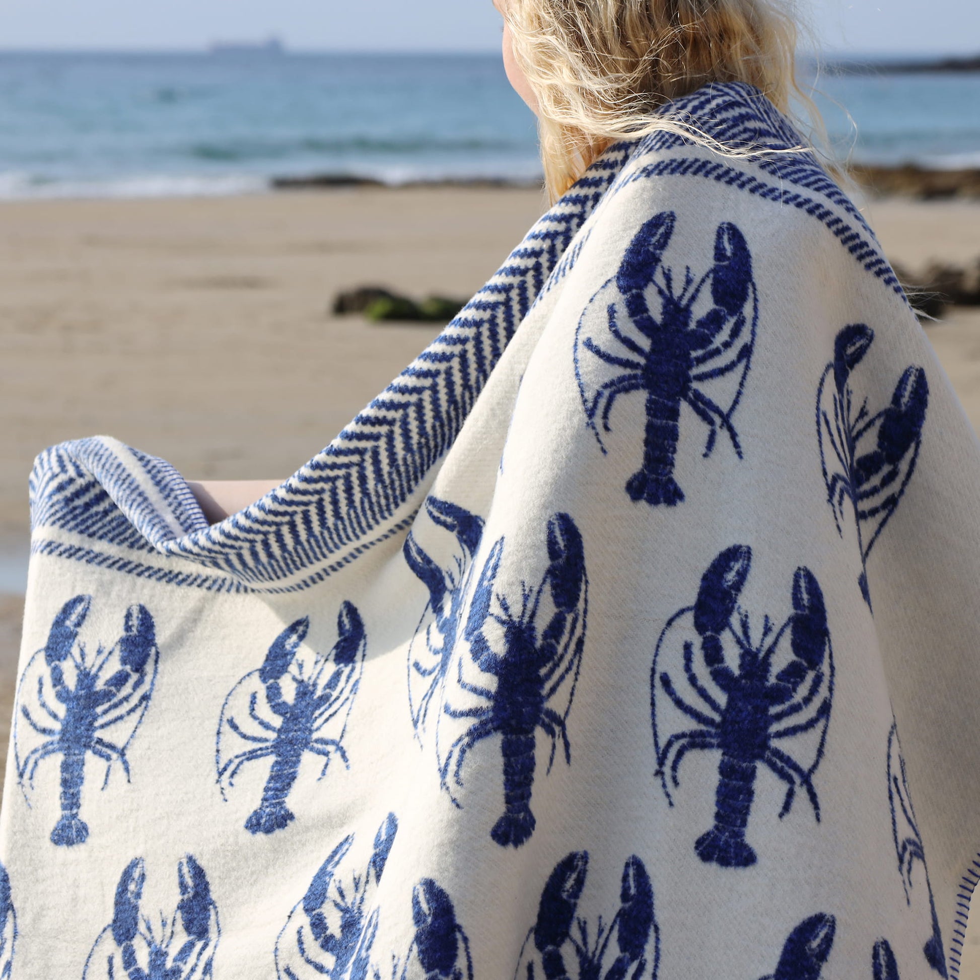 Person wearing a white blanket with blue lobster patterns on a beach.