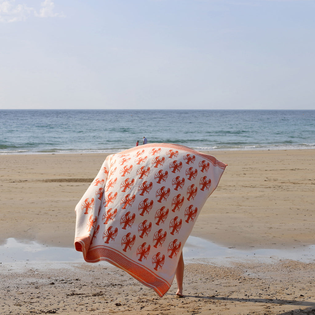 Person holding a beach towel with lobster pattern on a sandy beach.