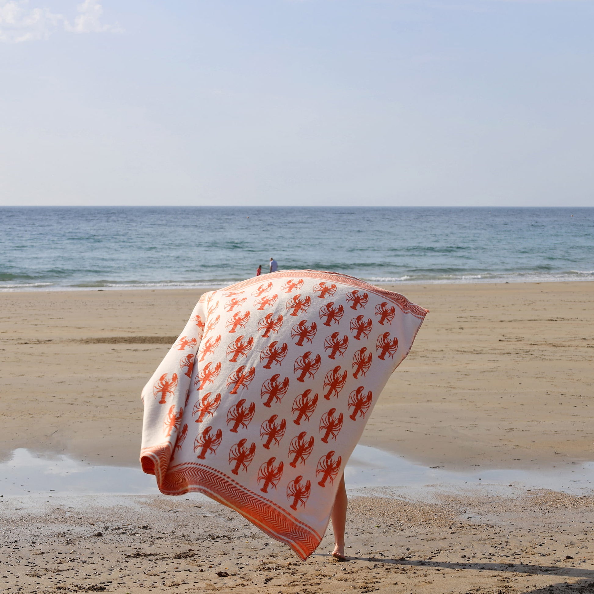 Person holding a beach towel with lobster pattern on a sandy beach.