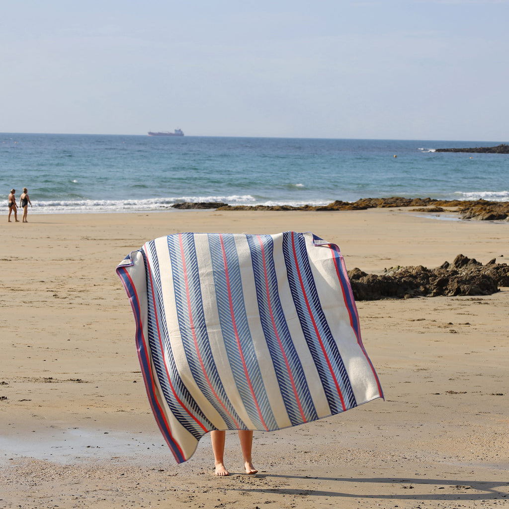 Person holding a striped beach towel on a sandy beach with ocean and sky in the background