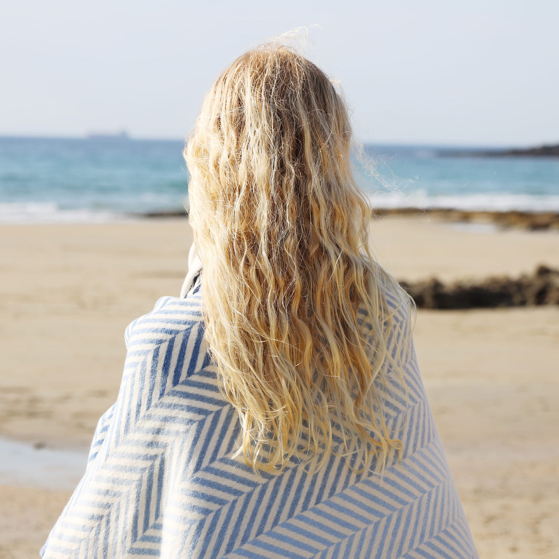Person with long blonde hair wrapped in a blue and white striped towel on a beach.