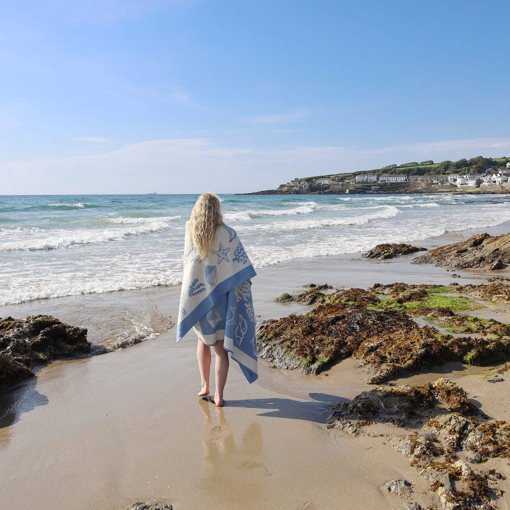 Person standing on a beach holding a blue towel, looking out at the ocean.