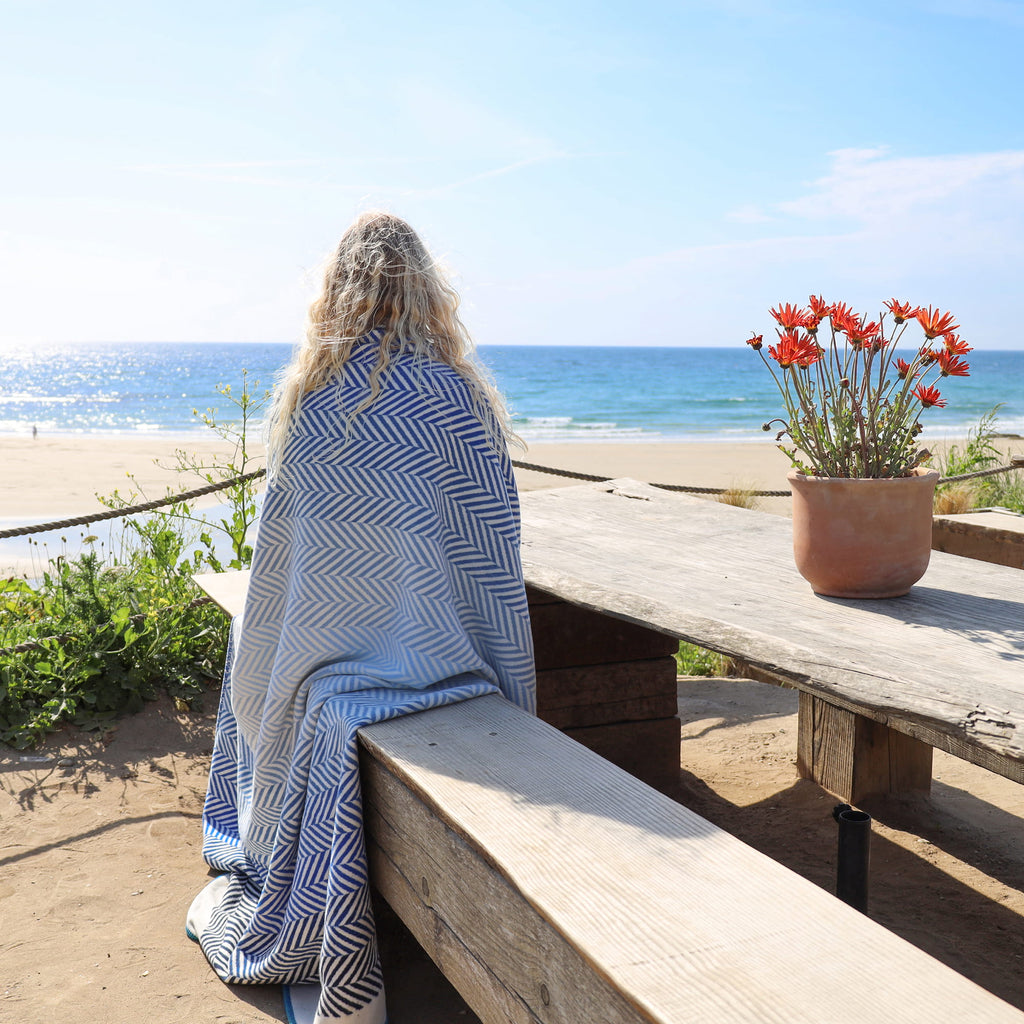 Person wrapped in a blue and white patterned towel sitting on a wooden bench by the ocean.