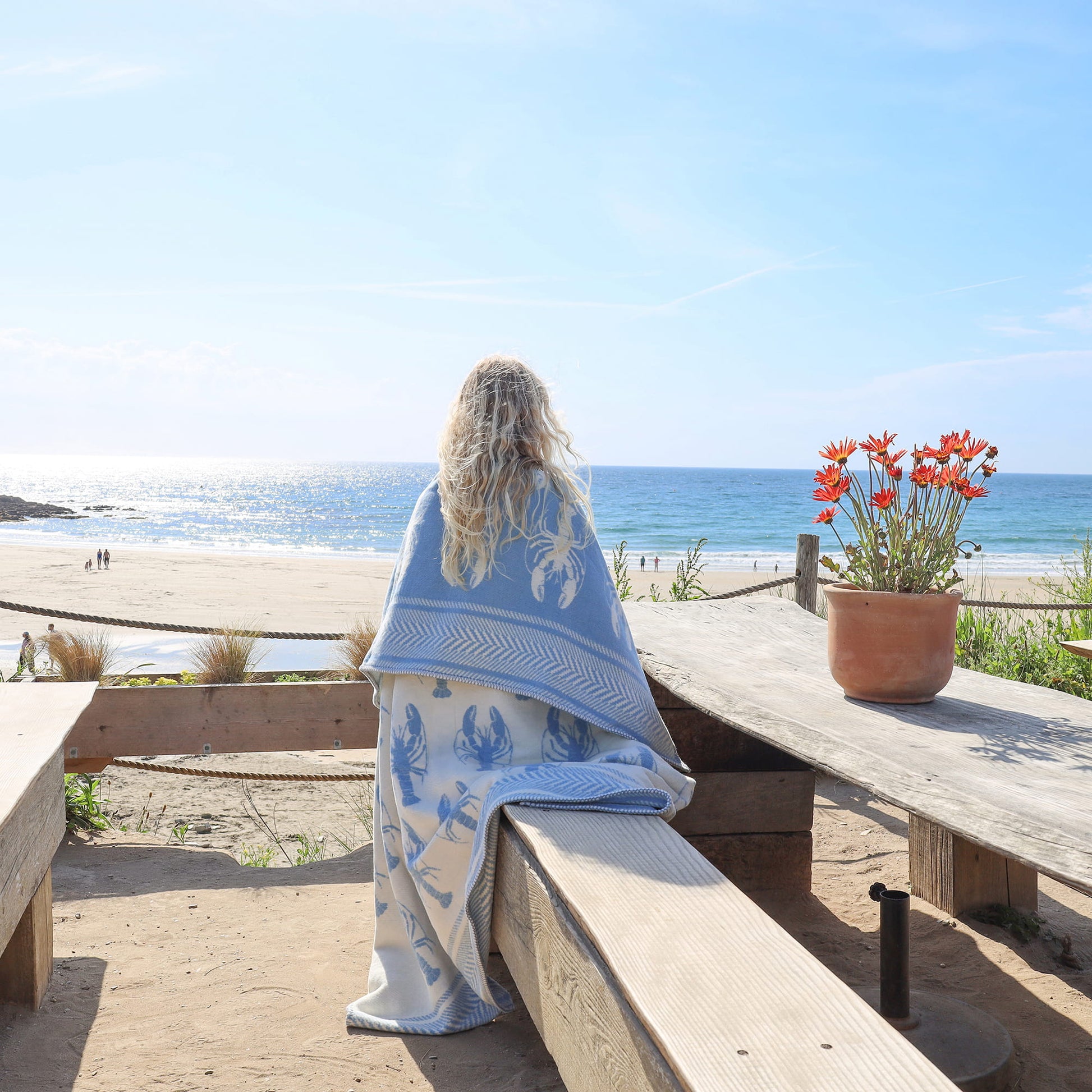 Person sitting on a wooden bench by the ocean, wrapped in a blue towel with a floral pattern, next to a potted plant.
