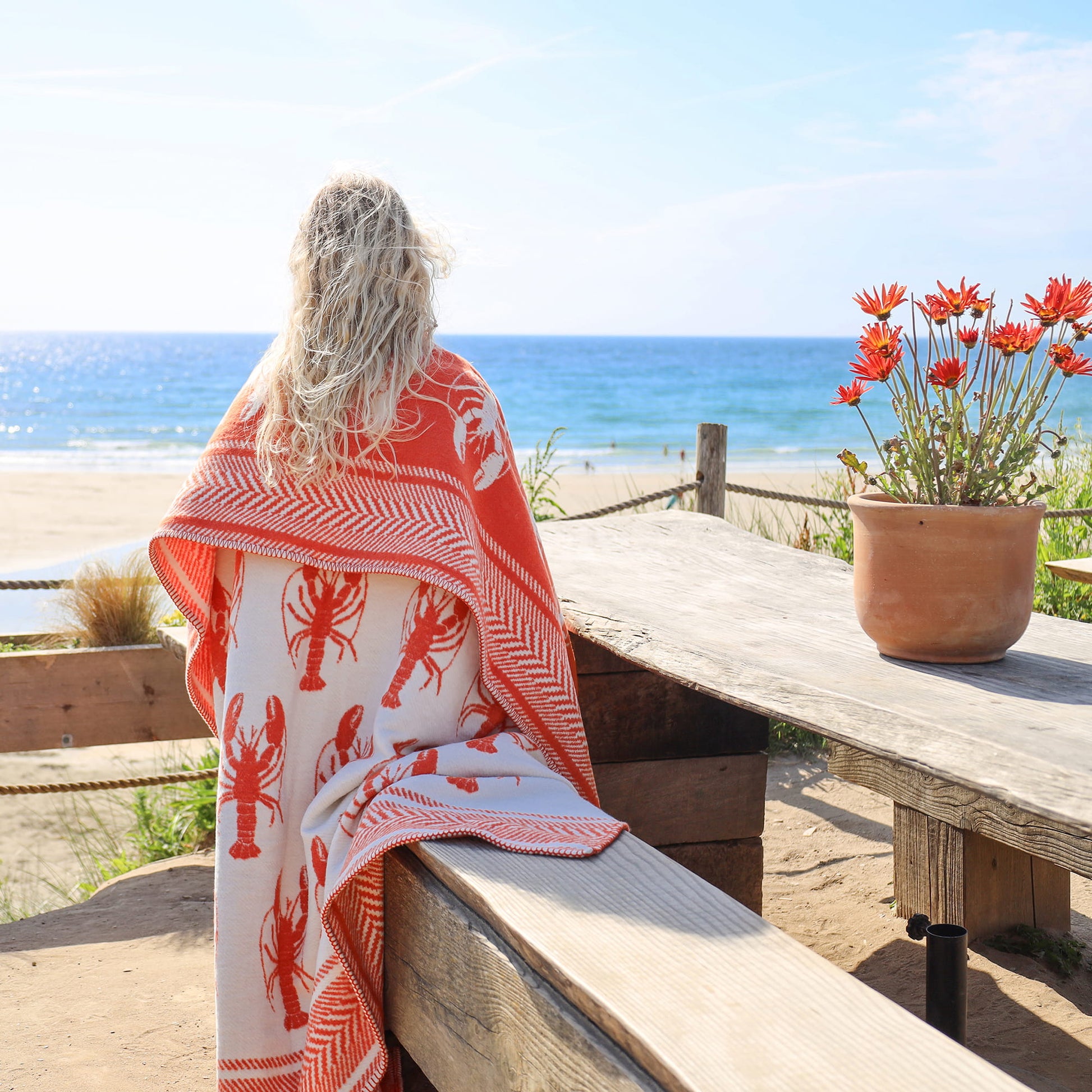 Person wrapped in a red and white patterned towel sitting on a wooden bench by the ocean.
