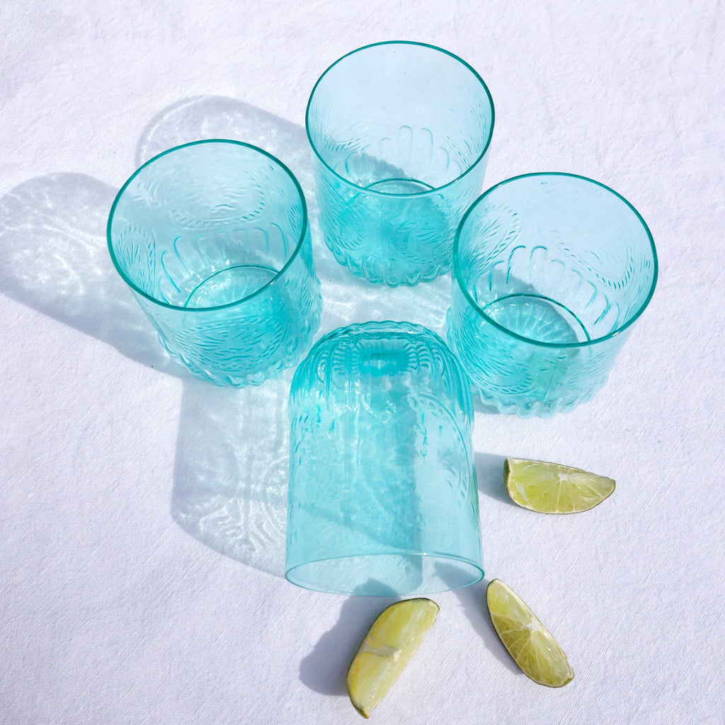 Set of four embossed turquoise glass tumblers arranged on a white surface with sunlight casting reflections, accompanied by lime wedges.