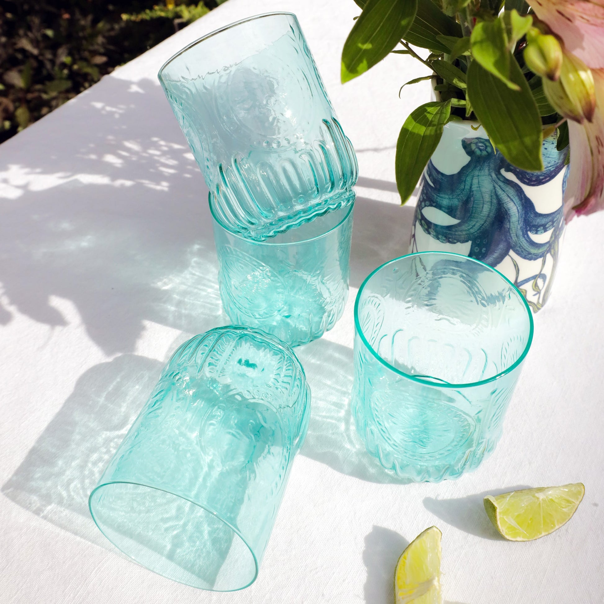 Set of turquoise glass tumblers on a white surface with a vase and lime slices in the background.