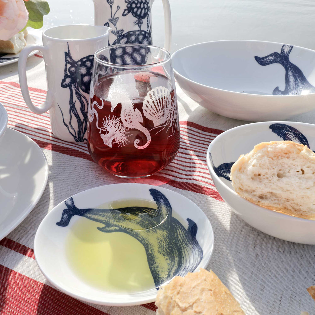 A set of white ceramic tableware with navy blue sea creature illustrations, including a bowl of bread, a plate with olive oil, a glass with red wine, and a mug, arranged on a striped table runner outdoors.