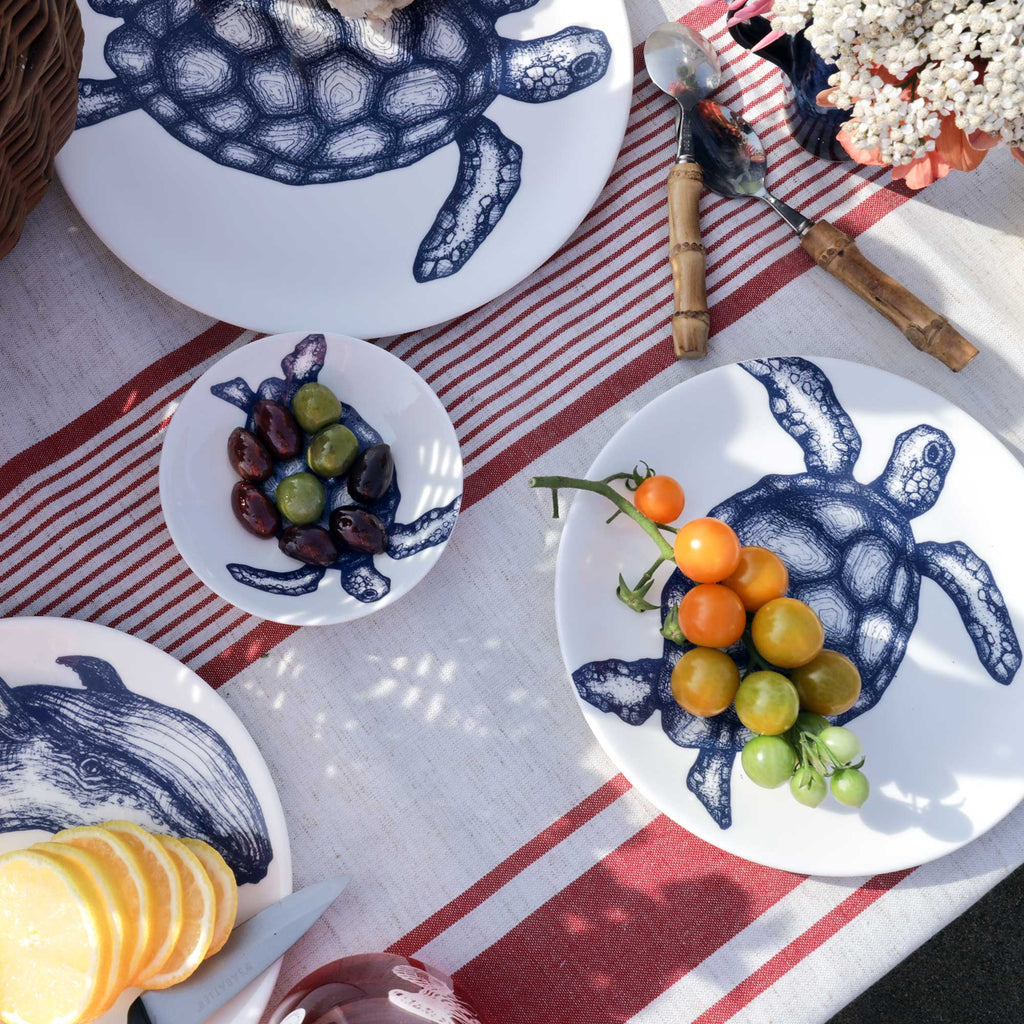 A styled flat-lay of a table setting featuring white ceramic plates and a bowl with a navy blue turtle illustration, set on a red and white striped runner with food props including cherry tomatoes, olives, and sliced lemons.
