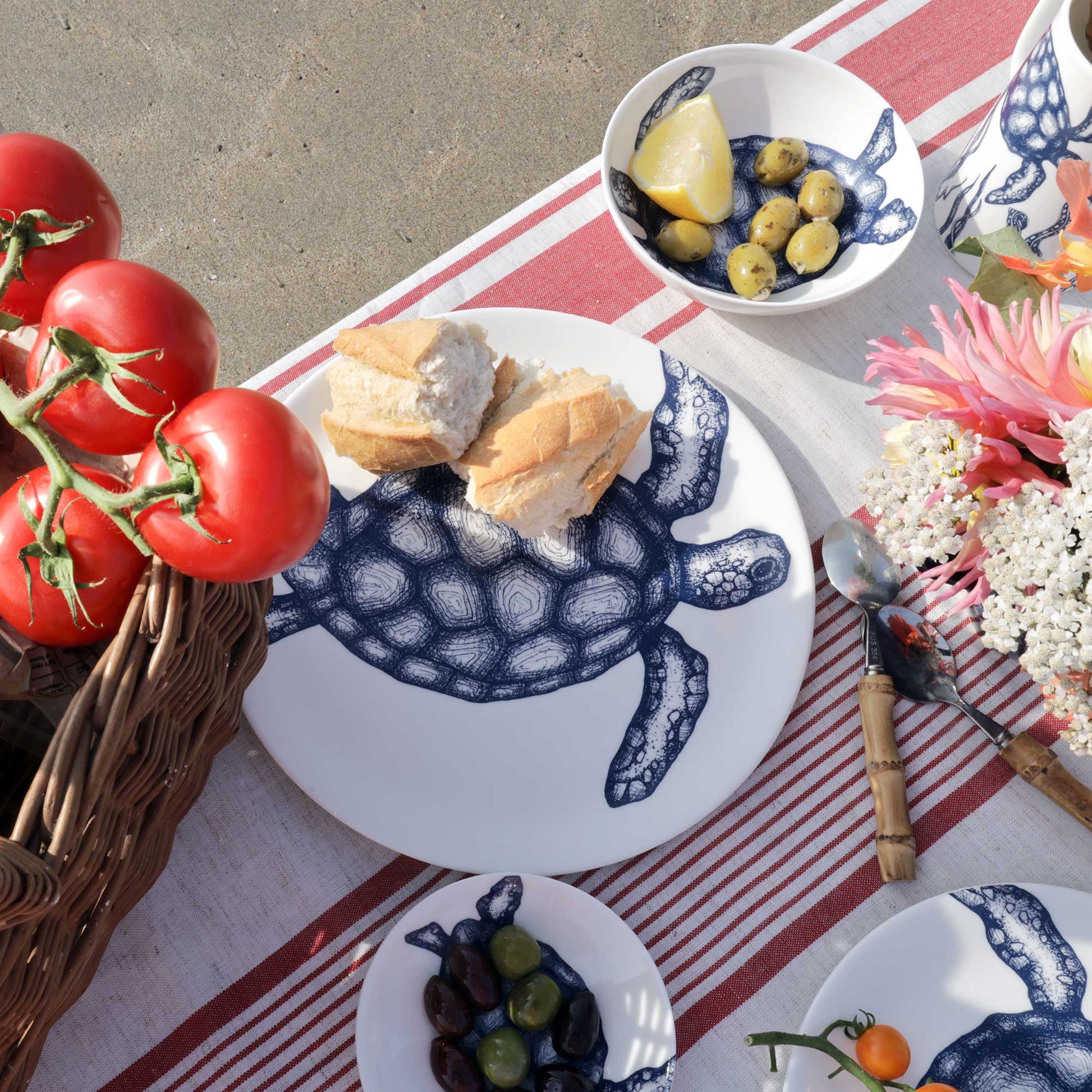 A styled outdoor picnic scene featuring a white ceramic plate with a navy blue turtle design, set on a red and white striped tablecloth with bread, olives, tomatoes, flowers, and cutlery.