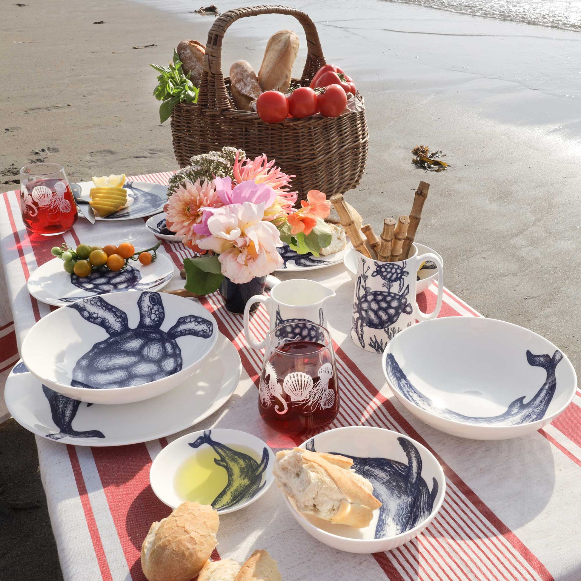 A styled outdoor dining setup on a beach tablecloth with white ceramic dishes featuring navy blue sea turtle illustrations, bread, fruit, flowers, and a wicker basket in the background.