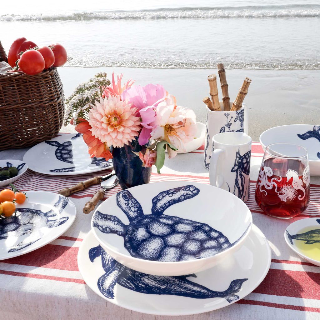 A styled outdoor dining table set with white ceramic tableware featuring a navy blue turtle print, placed on a red-and-white striped tablecloth with flowers, fruit, and a beach shoreline in the background.