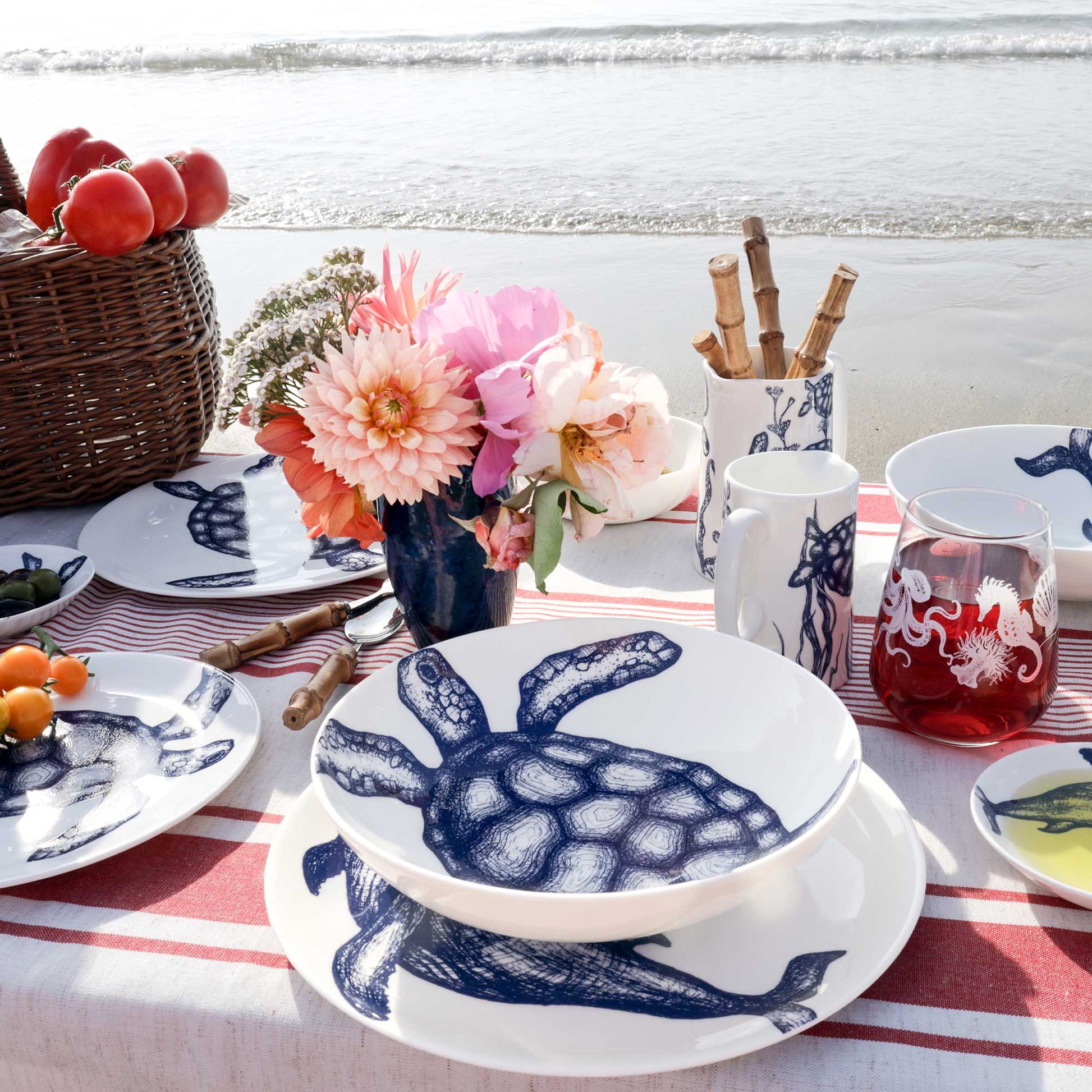 A styled outdoor dining table set with white ceramic tableware featuring a navy blue turtle print, placed on a red-and-white striped tablecloth with flowers, fruit, and a beach shoreline in the background.