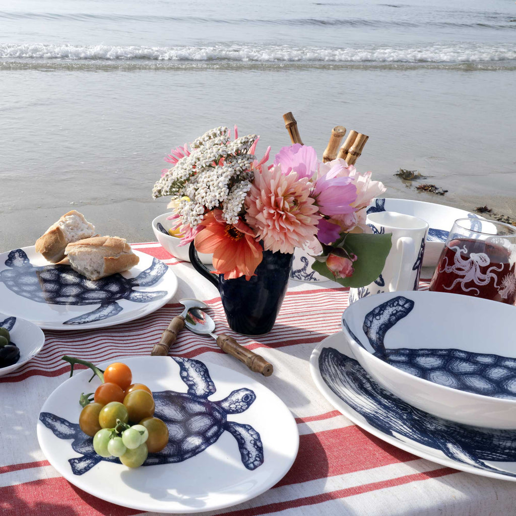 A styled beach picnic scene with white ceramic plates and bowls printed with a navy blue turtle motif, set on a red-and-white striped blanket with bread, fruit, a flower arrangement, and a mug in the foreground. The ocean and shoreline are visible in the background.