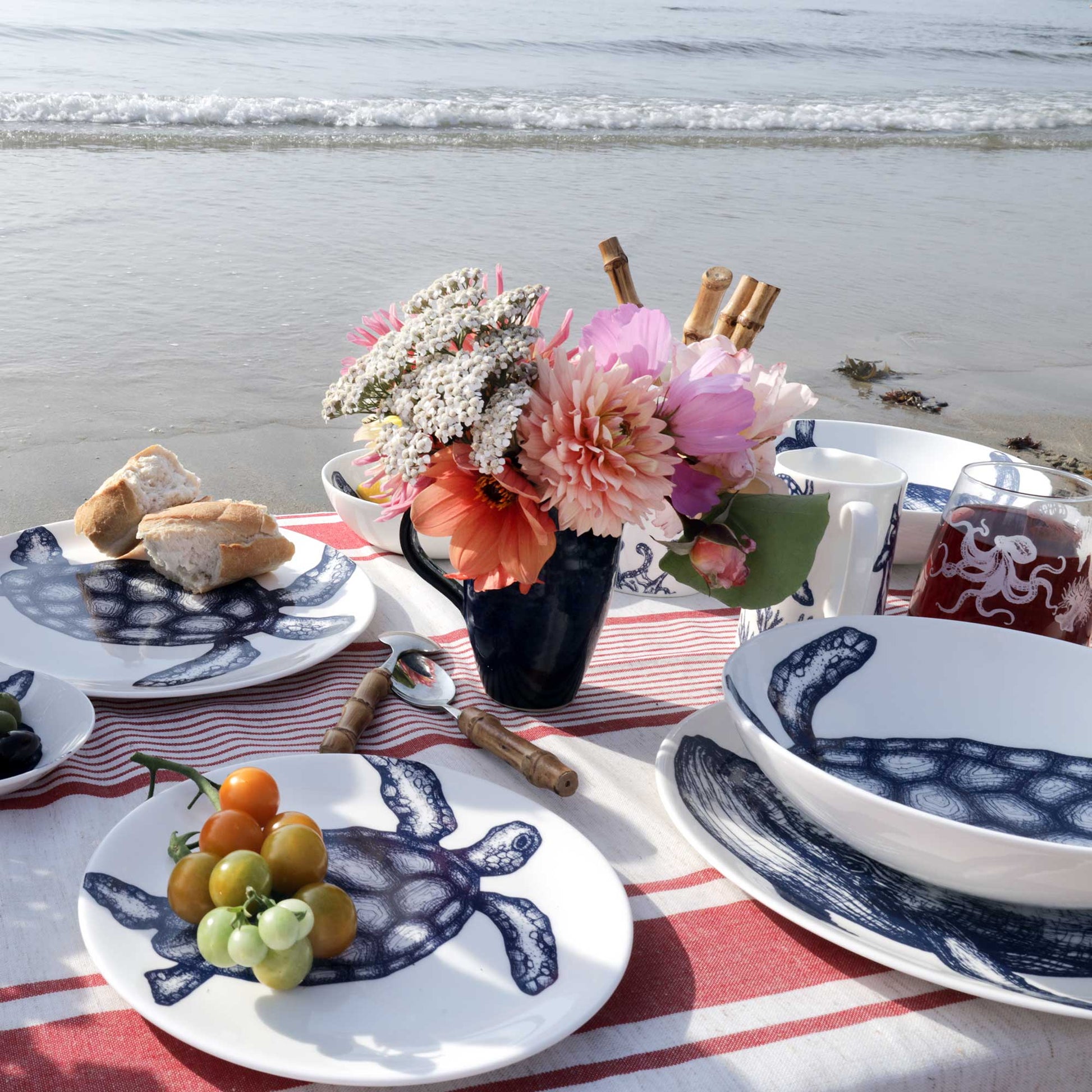 A styled beach picnic scene with white ceramic plates and bowls printed with a navy blue turtle motif, set on a red-and-white striped blanket with bread, fruit, a flower arrangement, and a mug in the foreground. The ocean and shoreline are visible in the background.