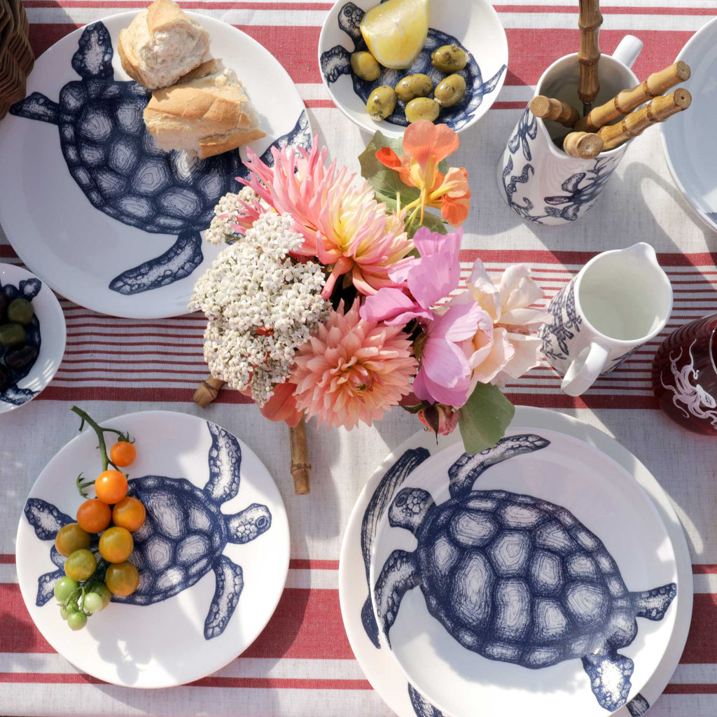 A styled picnic table setting featuring white ceramic plates with a navy blue turtle design, a red and white striped tablecloth, a floral centerpiece, and various food items including bread, olives, and cherry tomatoes.