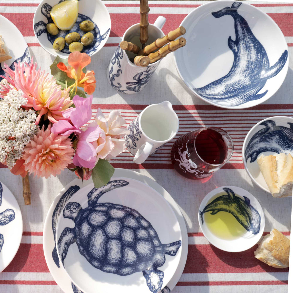 A styled picnic-style tabletop scene with white ceramic dishes and cups printed in navy blue sea creatures, including a whale, turtle, octopus, and fish. The table is set with a red-and-white striped cloth, flowers, olives, bread, and a drink.