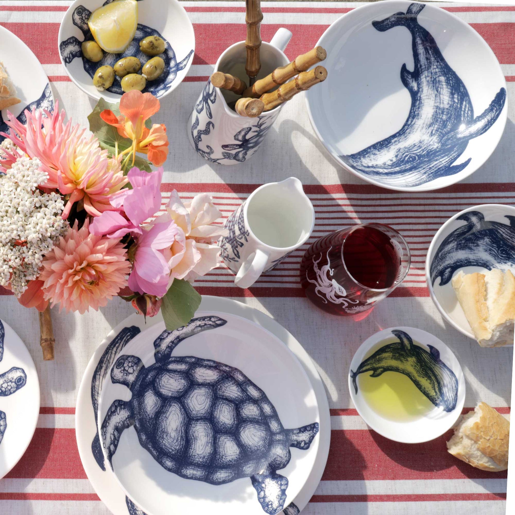 A styled picnic-style tabletop scene with white ceramic dishes and cups printed in navy blue sea creatures, including a whale, turtle, octopus, and fish. The table is set with a red-and-white striped cloth, flowers, olives, bread, and a drink.