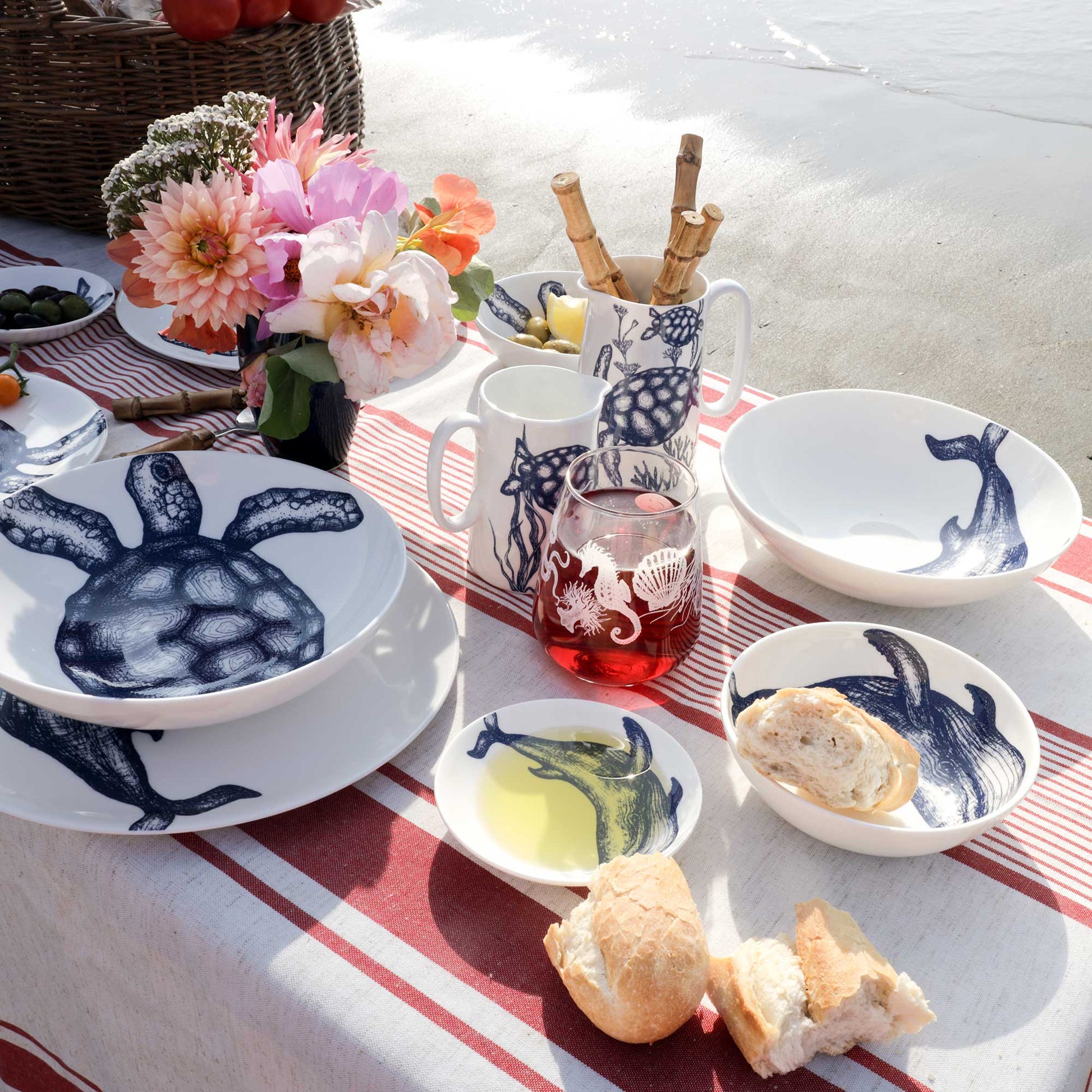 A styled outdoor dining table set with white ceramic dishes and mugs decorated with navy blue sea turtle and whale illustrations, alongside bread, flowers, and a drink, on a red-and-white striped blanket with sand in the background.