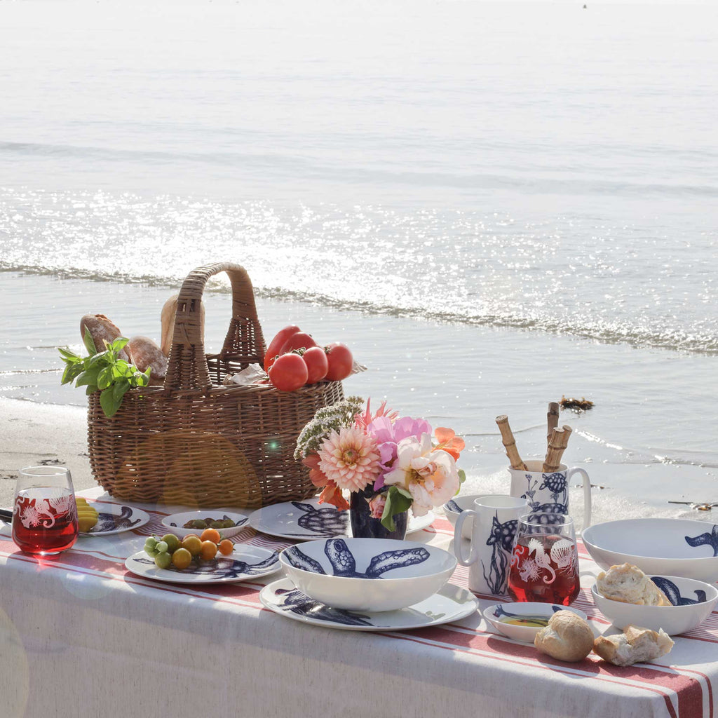 A styled beach picnic setup on a white tablecloth with blue-and-white ceramic dishes, a wicker basket of bread and fruit, flowers, and glassware, set against a sunny ocean shoreline.