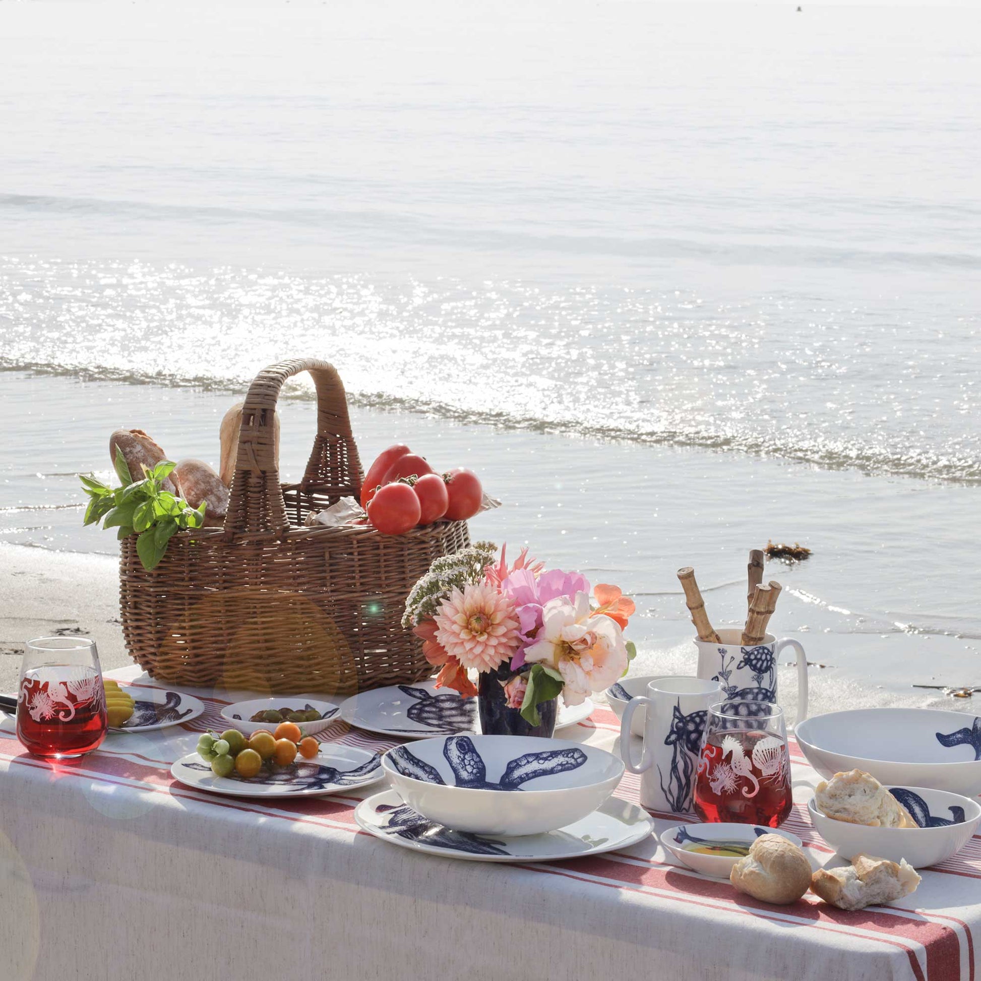 A styled beach picnic setup on a white tablecloth with blue-and-white ceramic dishes, a wicker basket of bread and fruit, flowers, and glassware, set against a sunny ocean shoreline.