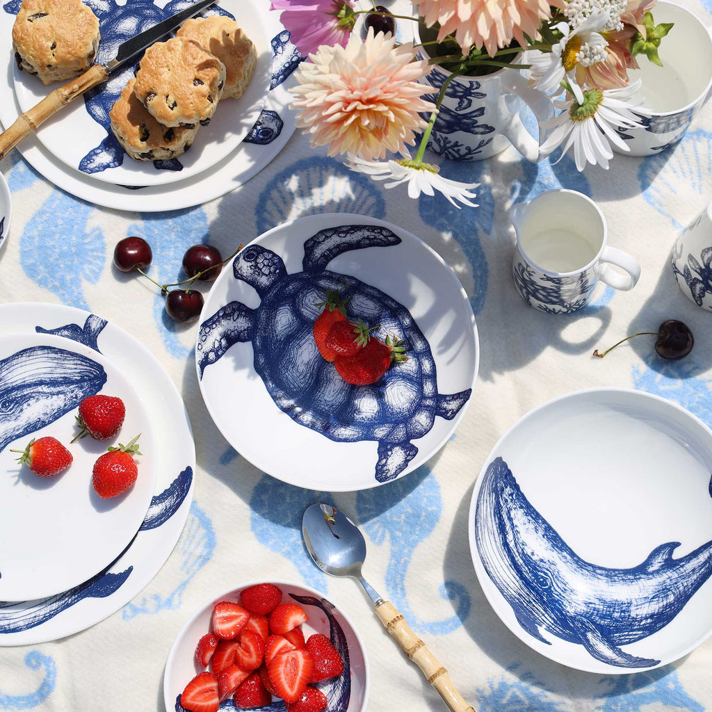A styled tabletop flat lay of a blue and white ceramic tableware set, featuring plates and bowls with blue sea creature illustrations like turtles, whales, and fish. The set is arranged on a blue patterned tablecloth with strawberries, biscuits, flowers, and a teapot.