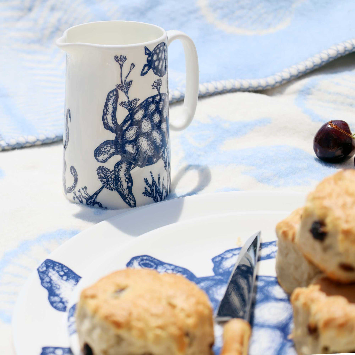 A white ceramic pitcher with a blue sea turtle and marine life illustration, set on a blue and white patterned blanket next to a plate of scones and a knife.